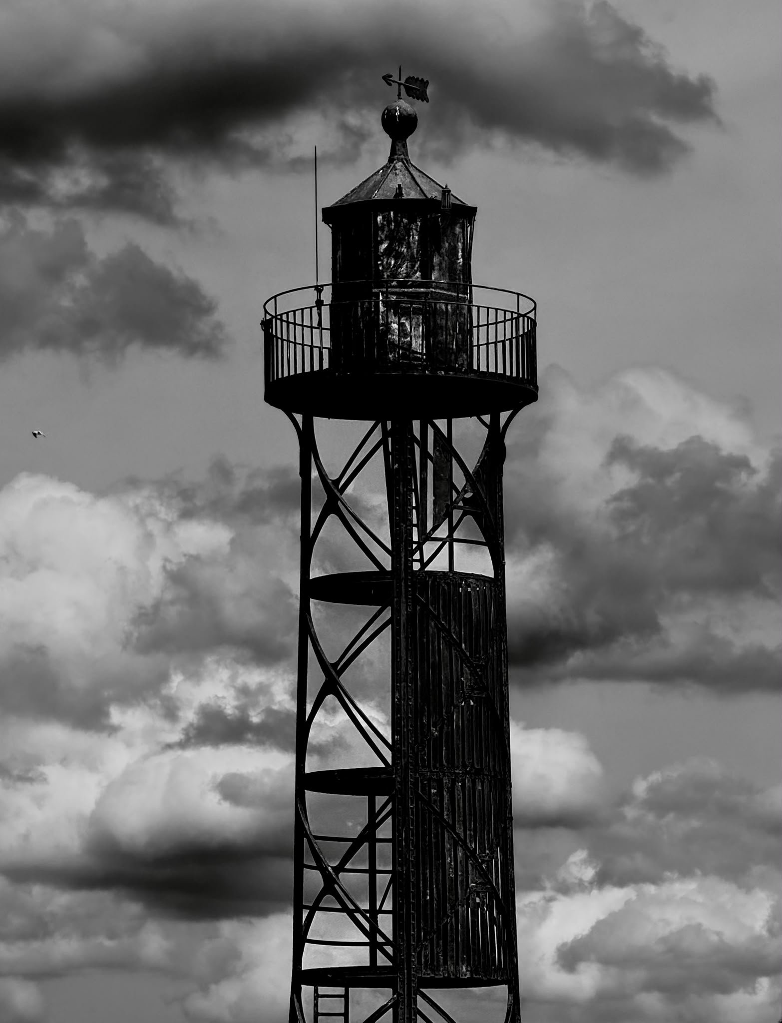 A black and white photo of a tall lighthouse with a spiral staircase and a weather vane on top, against a cloudy sky.