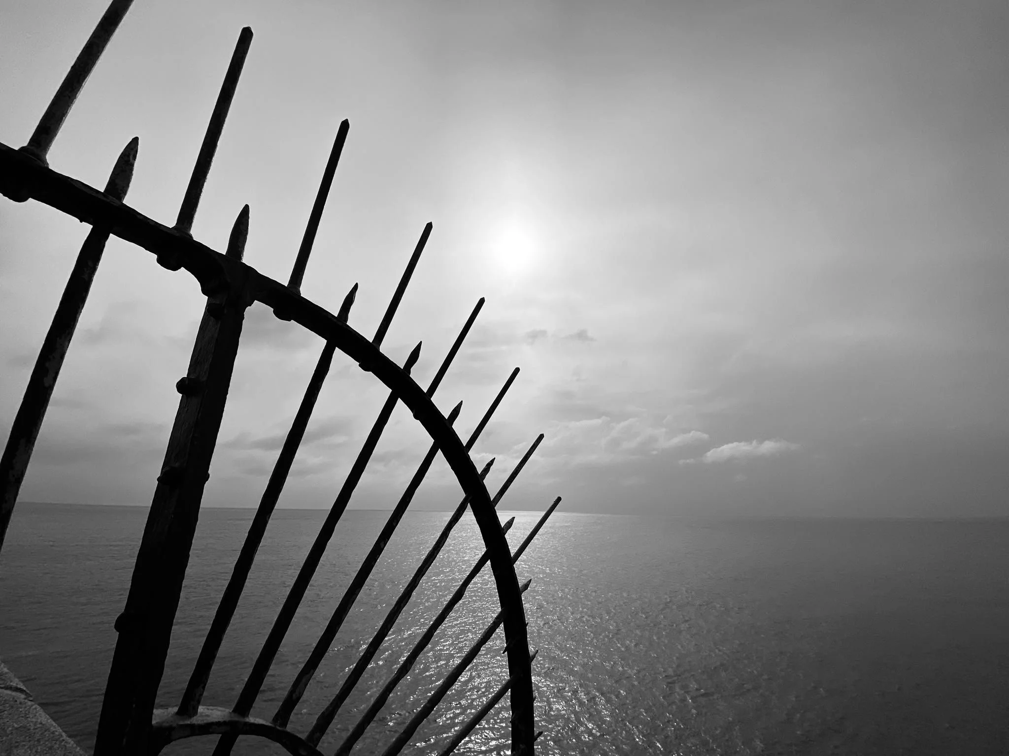 Black and white photograph of a weathered metal fence in the foreground, with the ocean and a cloudy sky in the background.