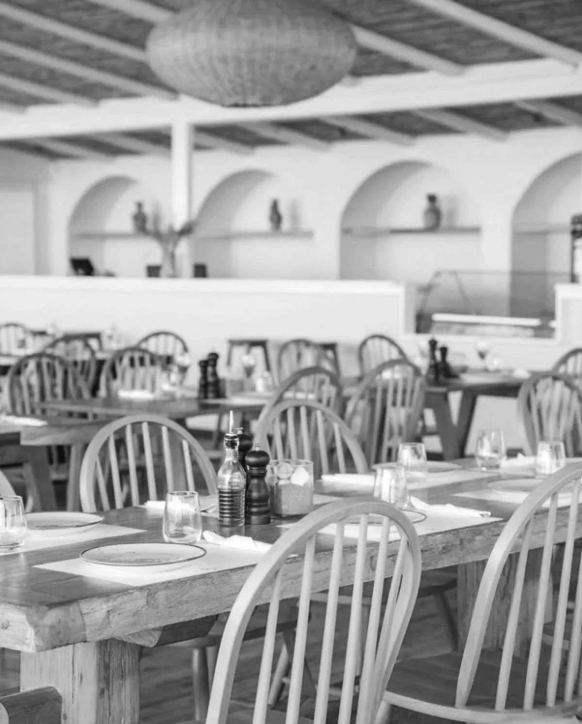 A black and white photo of an empty restaurant dining area with wooden tables, chairs, and various condiments on the tables, including salt and pepper shakers, and bottles of oil or vinegar. In the background, there are spaced alcoves with decorative vases and a ceiling with wooden beams and a round paper lantern.