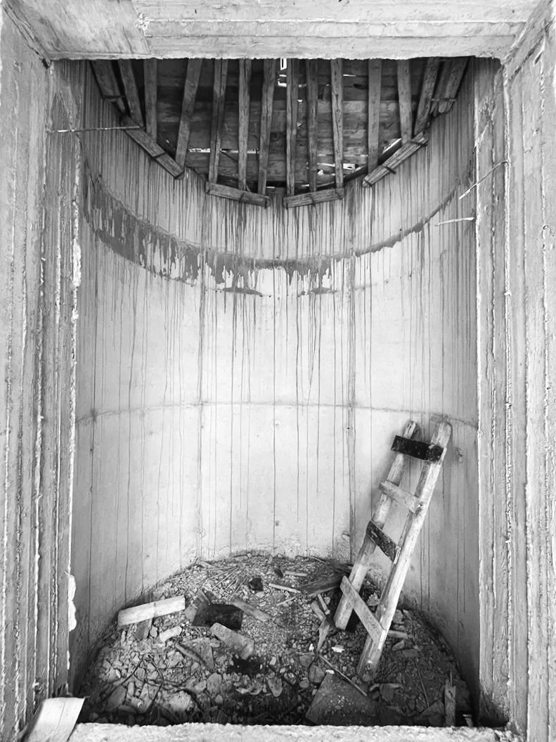 Black and white photo of an unfinished room or structure with a small, round, partially constructed space. The walls are plain with vertical paneling and stained water marks. A ladder leans against the far wall. The floor is filled with debris, bricks, and scrap materials, indicating ongoing construction.