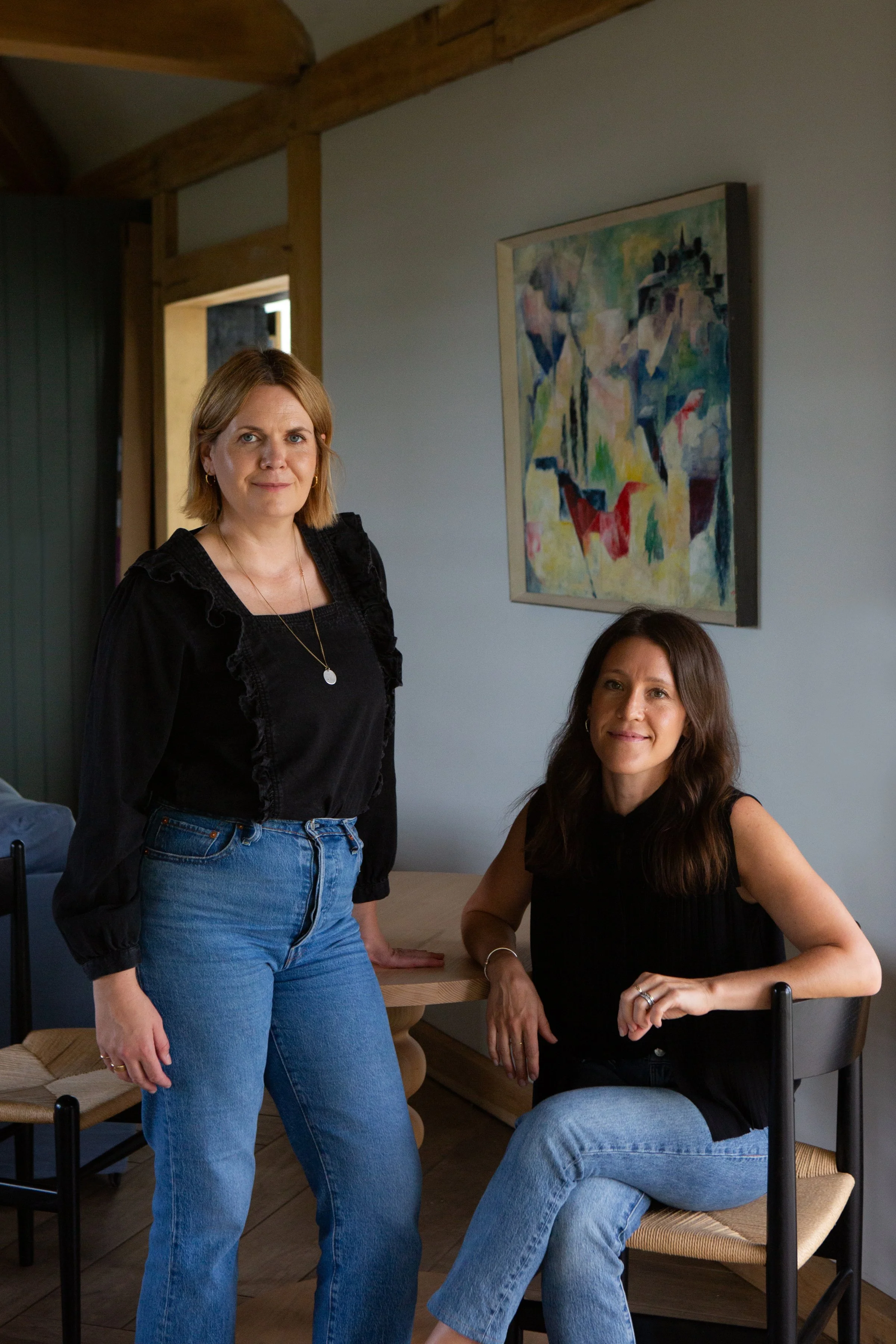 Two women in a room with wooden beams and an abstract painting on the wall. One woman is standing, wearing a black blouse and blue jeans, with shoulder-length blonde hair. The other woman is seated, wearing a black sleeveless top and blue jeans, with long dark hair. The room has a dining table and chairs.