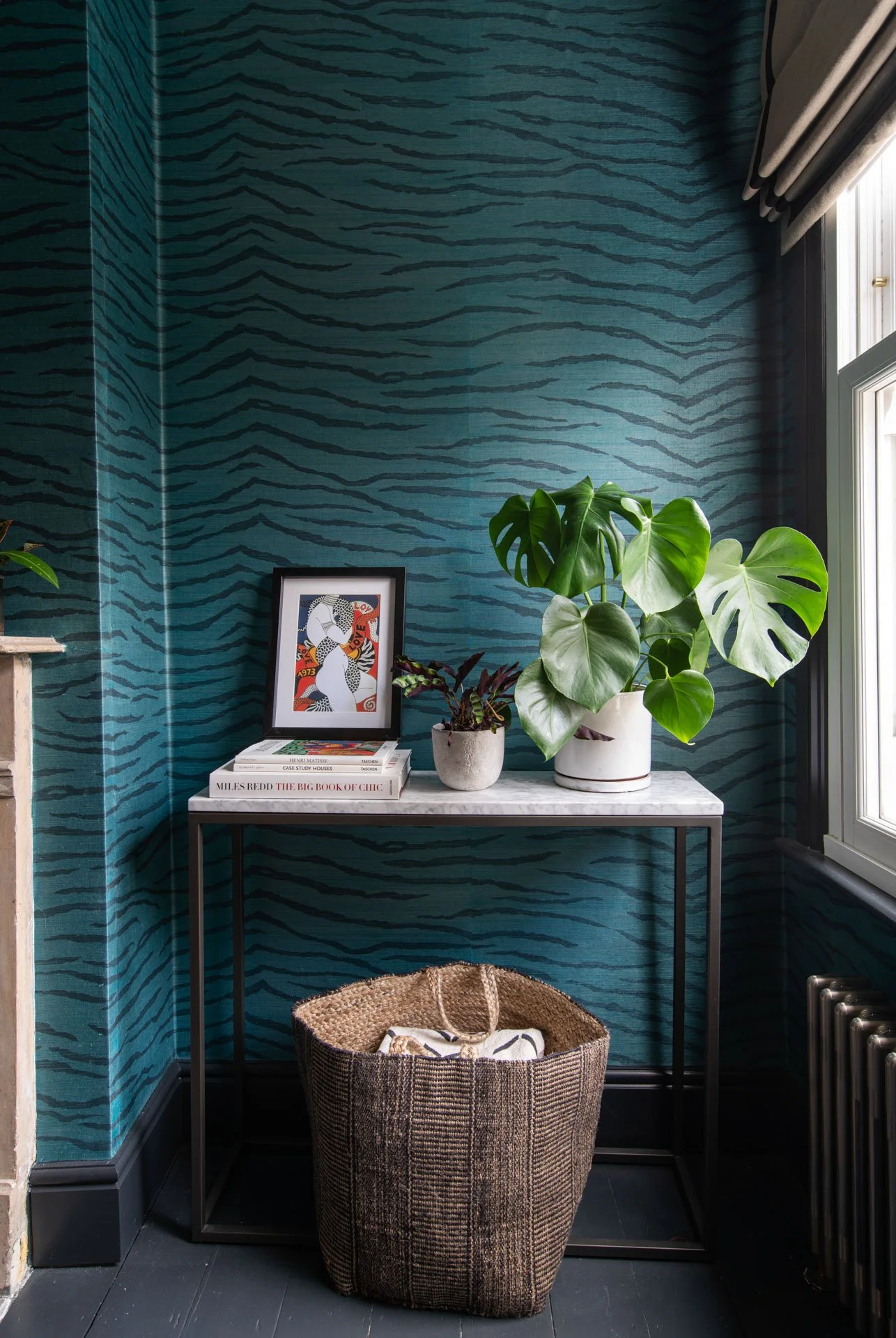 A corner of a room with teal animal print wallpaper, a black metal table with potted plants, a stack of books, and a framed art print. A woven basket with a blanket is on the dark floor, beside a window with gray curtains.