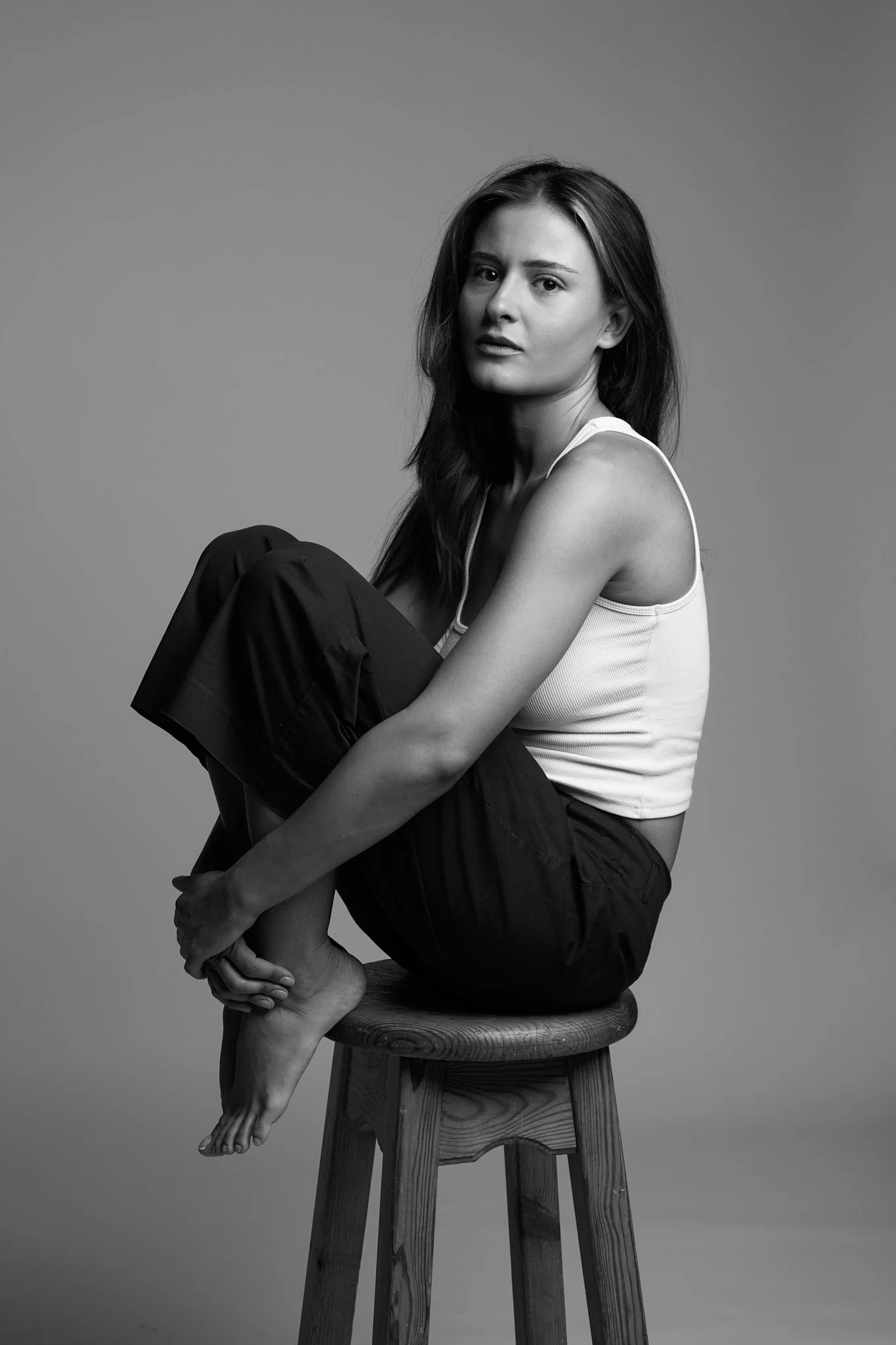 Black and white photo of a young woman sitting barefoot on a wooden stool, with her arms wrapped around her bent knees, looking at the camera with a neutral expression against a plain background.