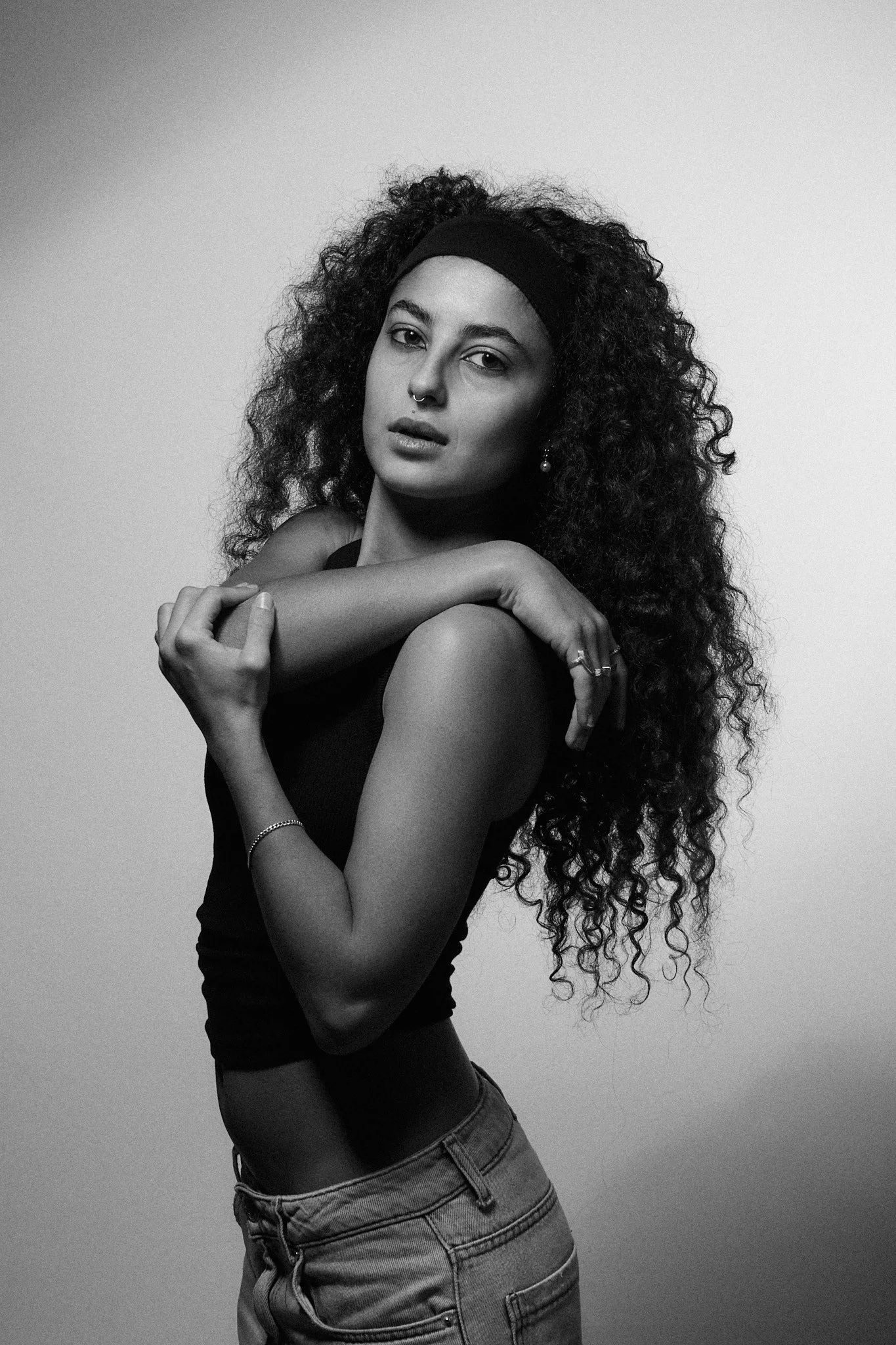 Black and white photo of a woman with curly hair wearing a headband, posing with one arm across her shoulders and looking at the camera.
