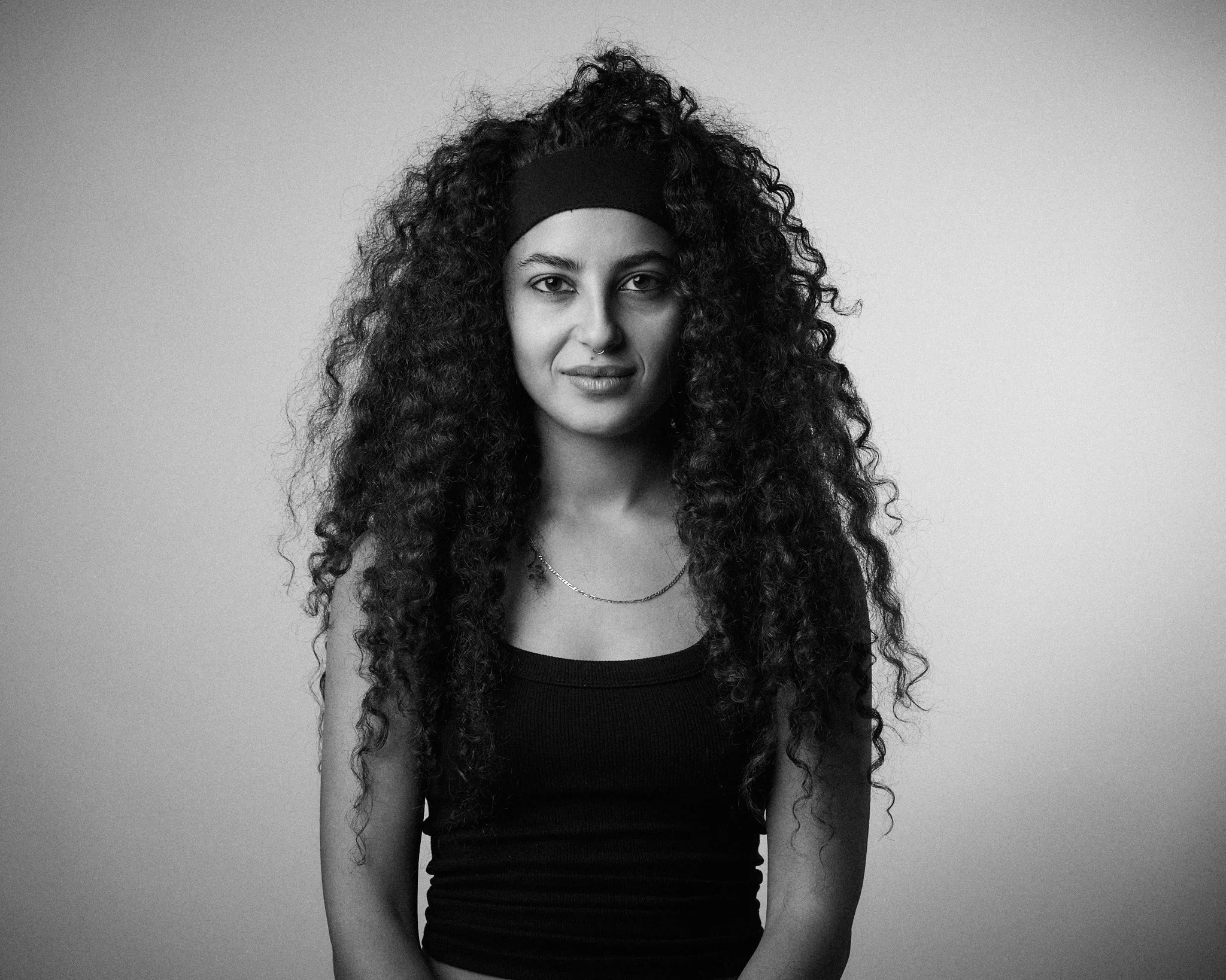 A woman with long, curly hair wearing a black headband and a black tank top, looking into the camera against a plain gray background.