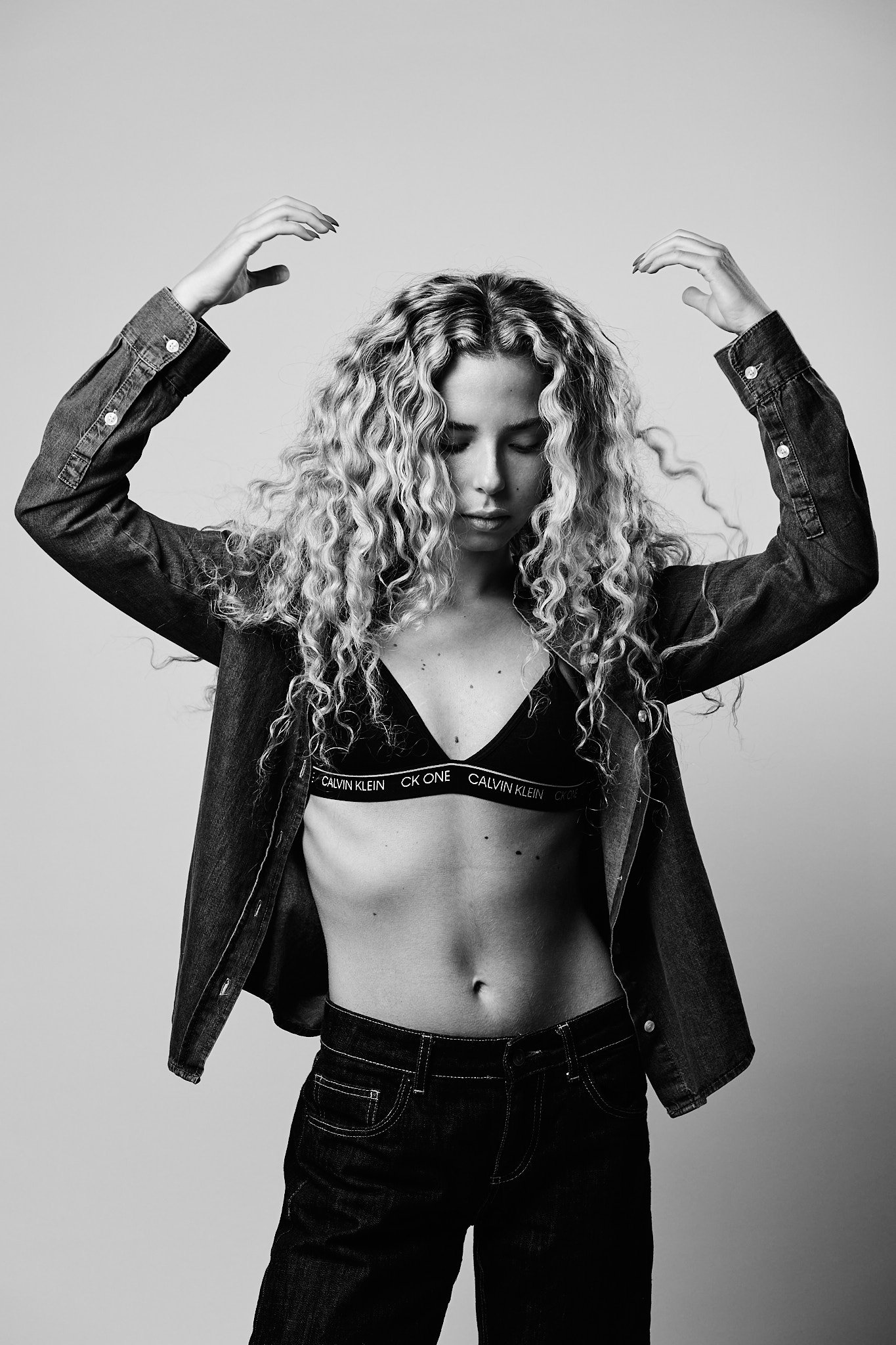 Black and white photo of a young woman with curly hair, wearing a Calvin Klein bra, a denim jacket, and jeans, with her eyes closed and arms raised above her head.