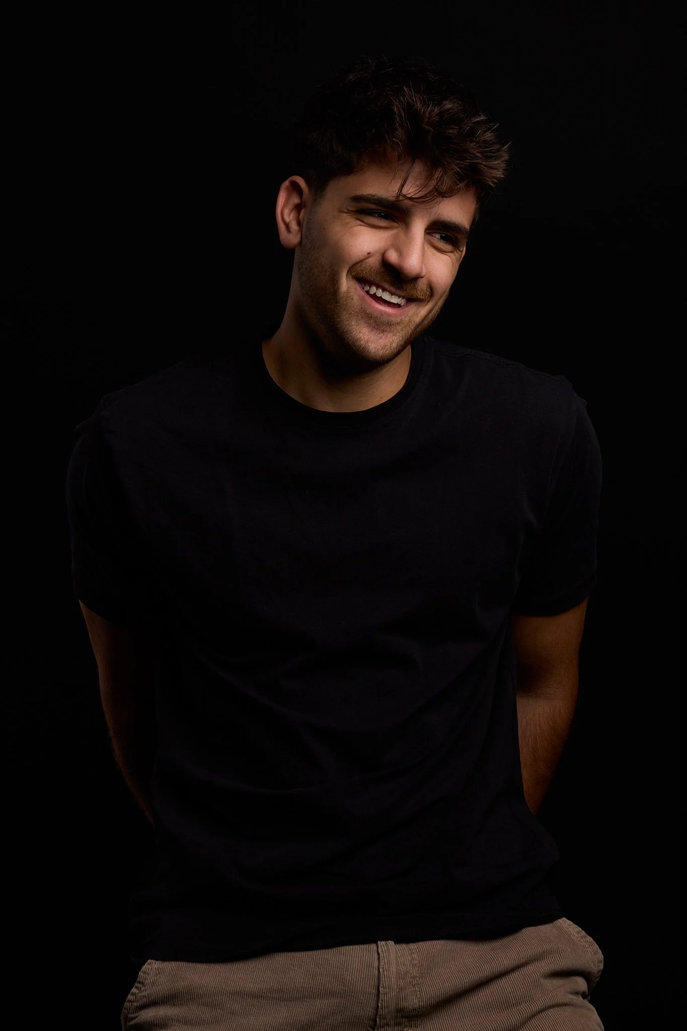 A young man with dark, curly hair and a beard is smiling and looking to his right. He is wearing a black t-shirt and khaki pants, standing against a black background.