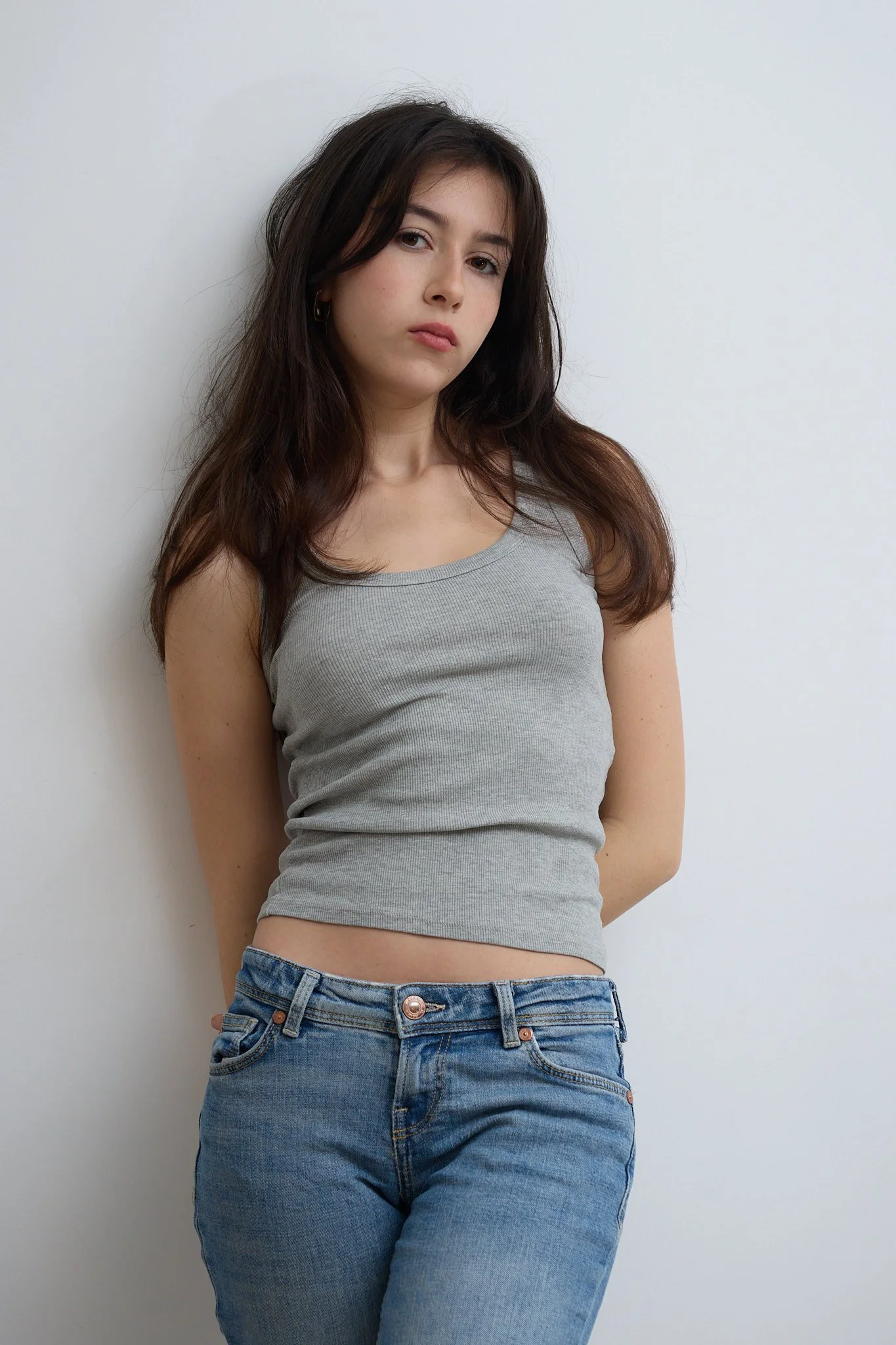 Young woman with dark brown hair, posing against a white wall, wearing a gray tank top and blue jeans.