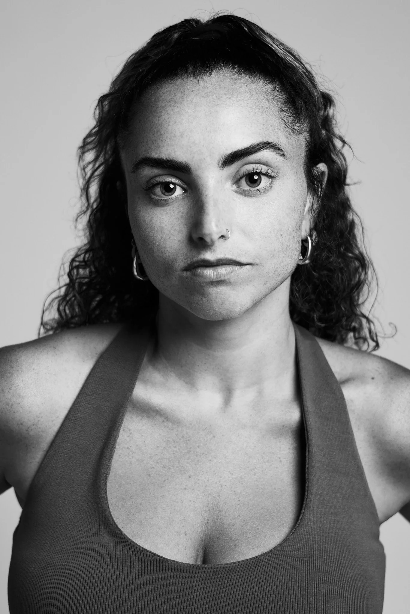 Black and white portrait of a woman with curly hair, hoop earrings, and a tank top, looking directly at the camera.