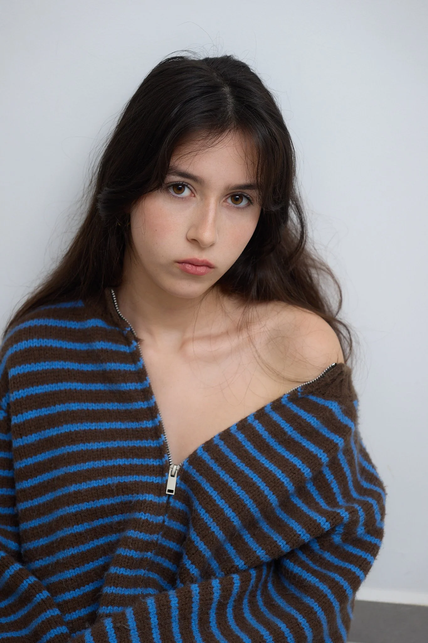 A young woman with long dark hair and light skin posing against a plain white wall. She is wearing a brown and blue striped zip-up sweater, which is partially unzipped, revealing her shoulder and collarbone.