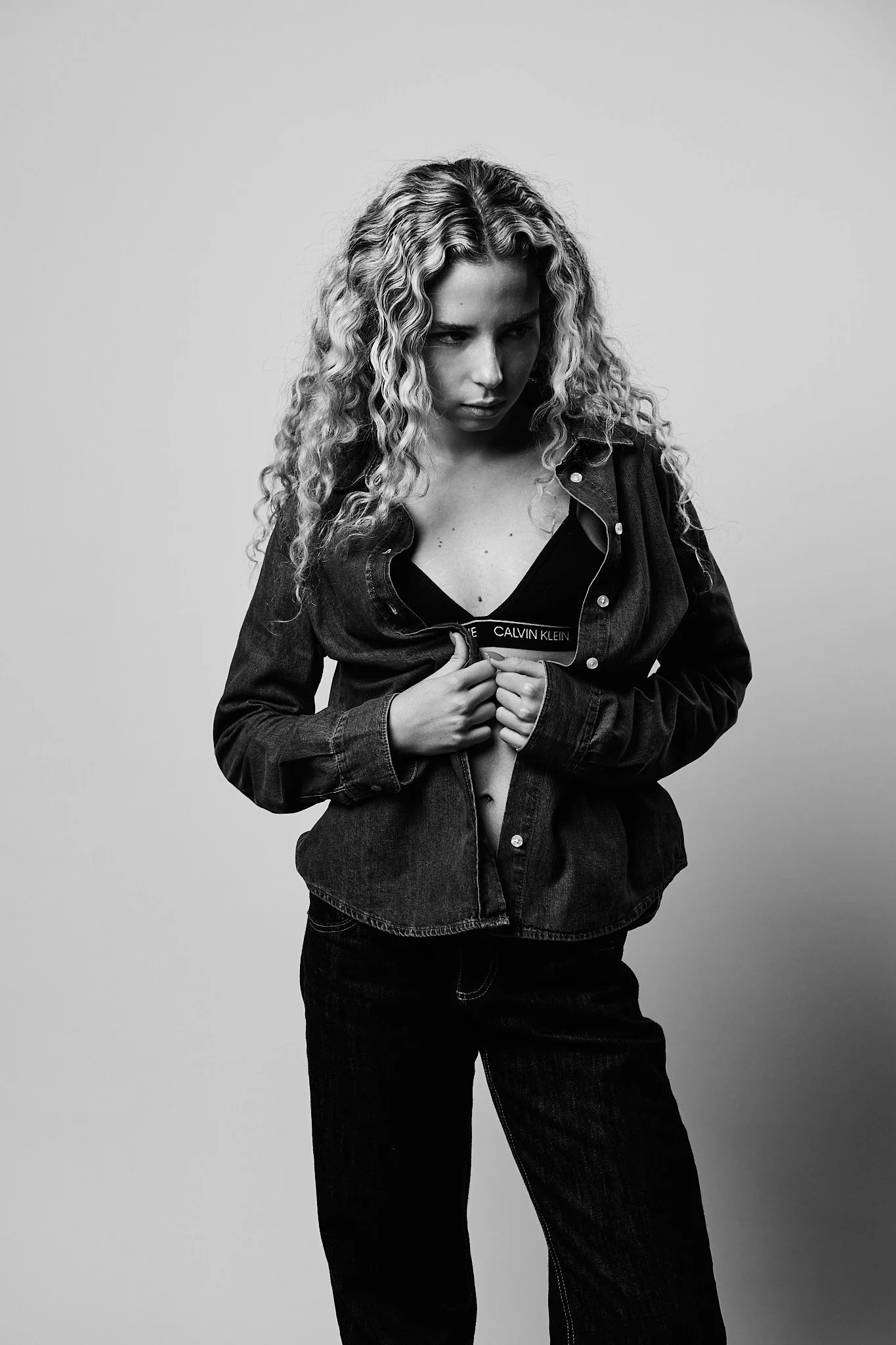 Black and white photo of a woman with curly hair wearing a partially unbuttoned denim jacket and a Calvin Klein bra, looking down with a serious expression.
