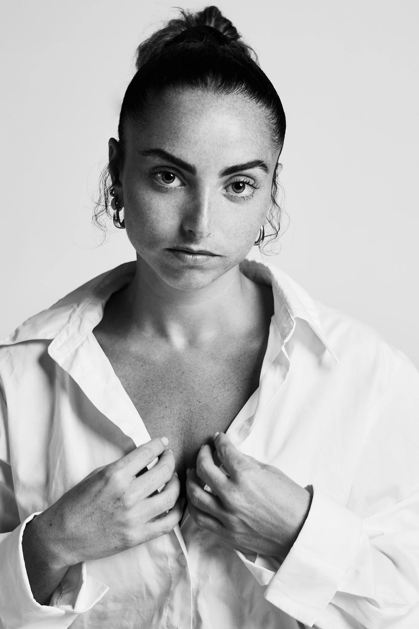 A black-and-white portrait of a woman with dark hair tied in a bun, wearing a white shirt, gazing directly at the camera, with her hands adjusting the collar of her shirt.
