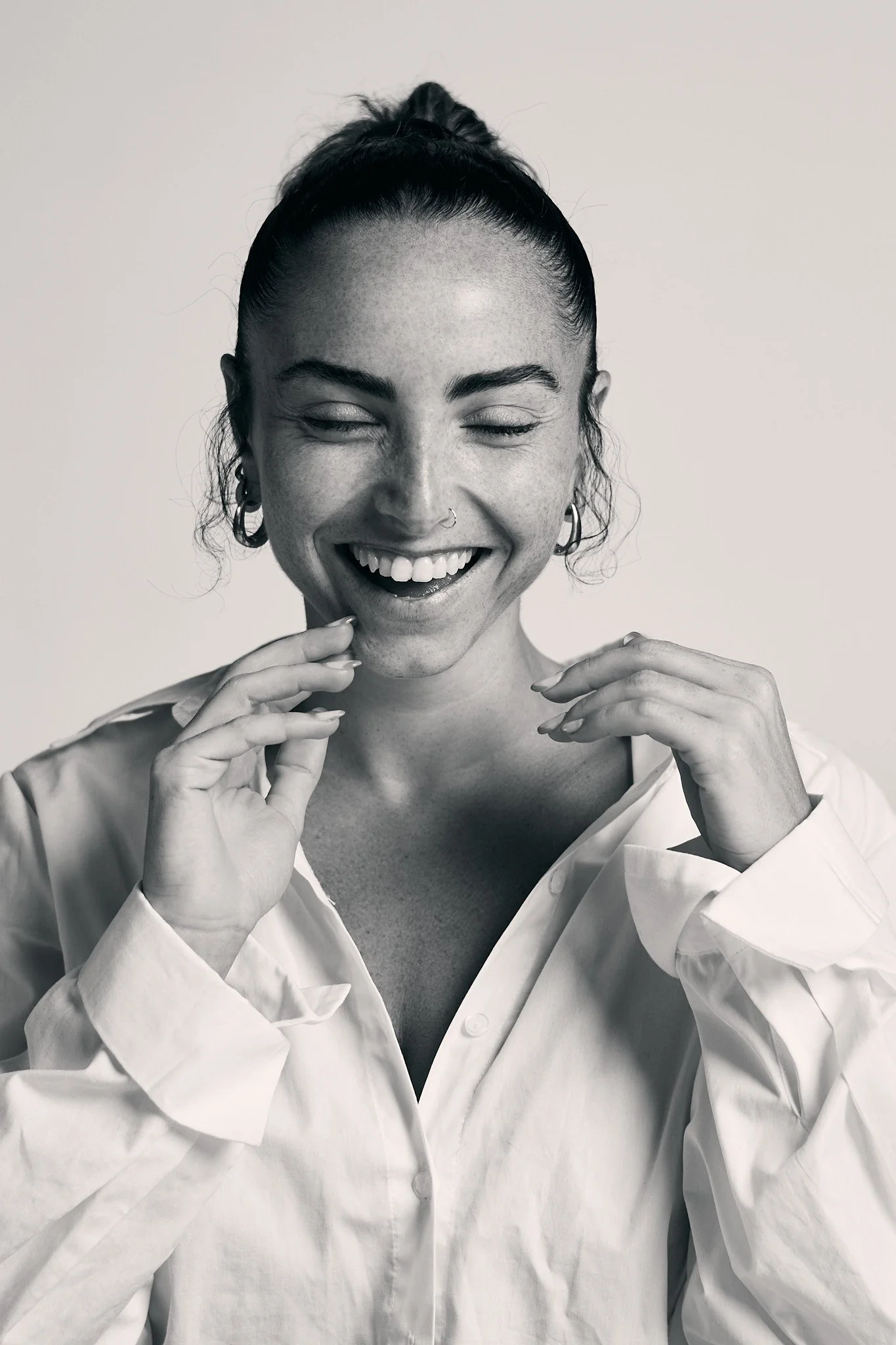 Black and white photo of a woman smiling with her eyes closed, wearing a white shirt with an open collar, and earrings.