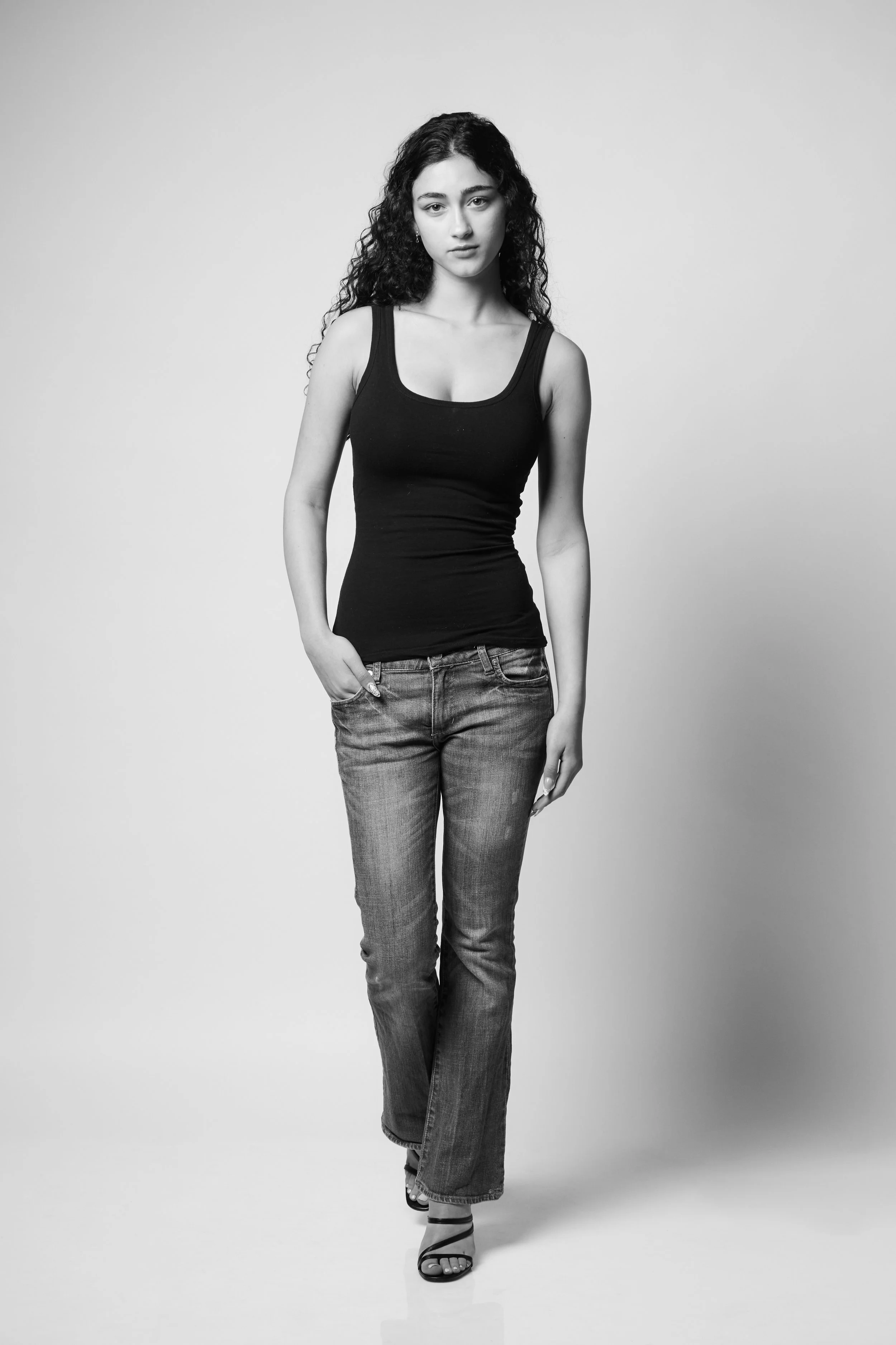 Black and white photo of a young woman with curly hair, wearing a black tank top, jeans, and sandals, standing with one hand in her pocket and looking at the camera.