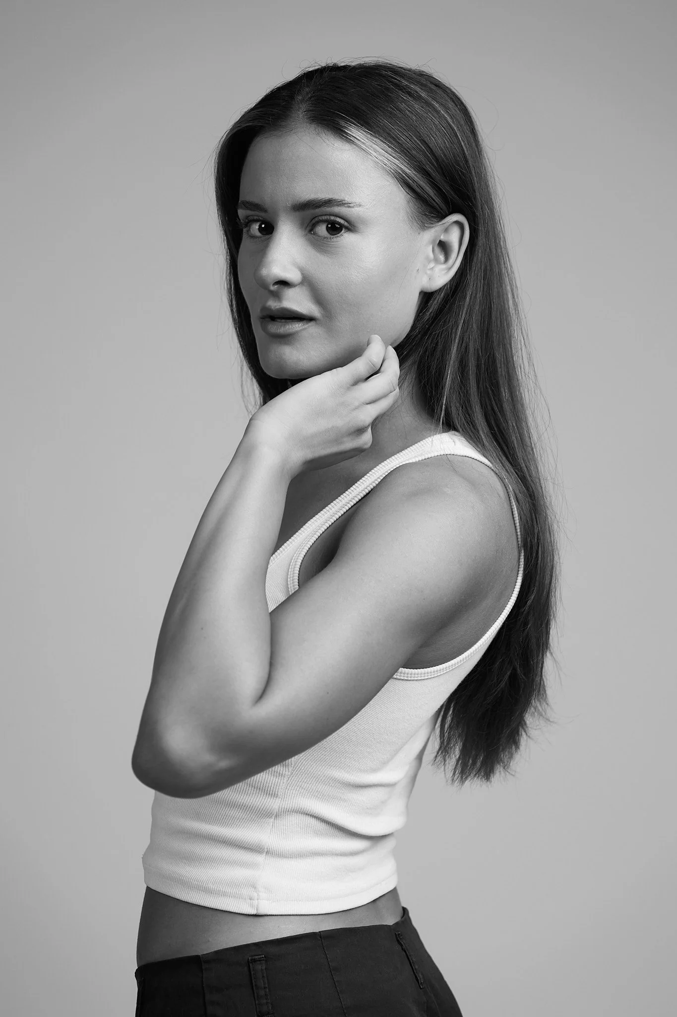 Black and white photo of a woman with long hair, wearing a sleeveless top and dark pants, posing with one hand gently touching her chin and looking at the camera.