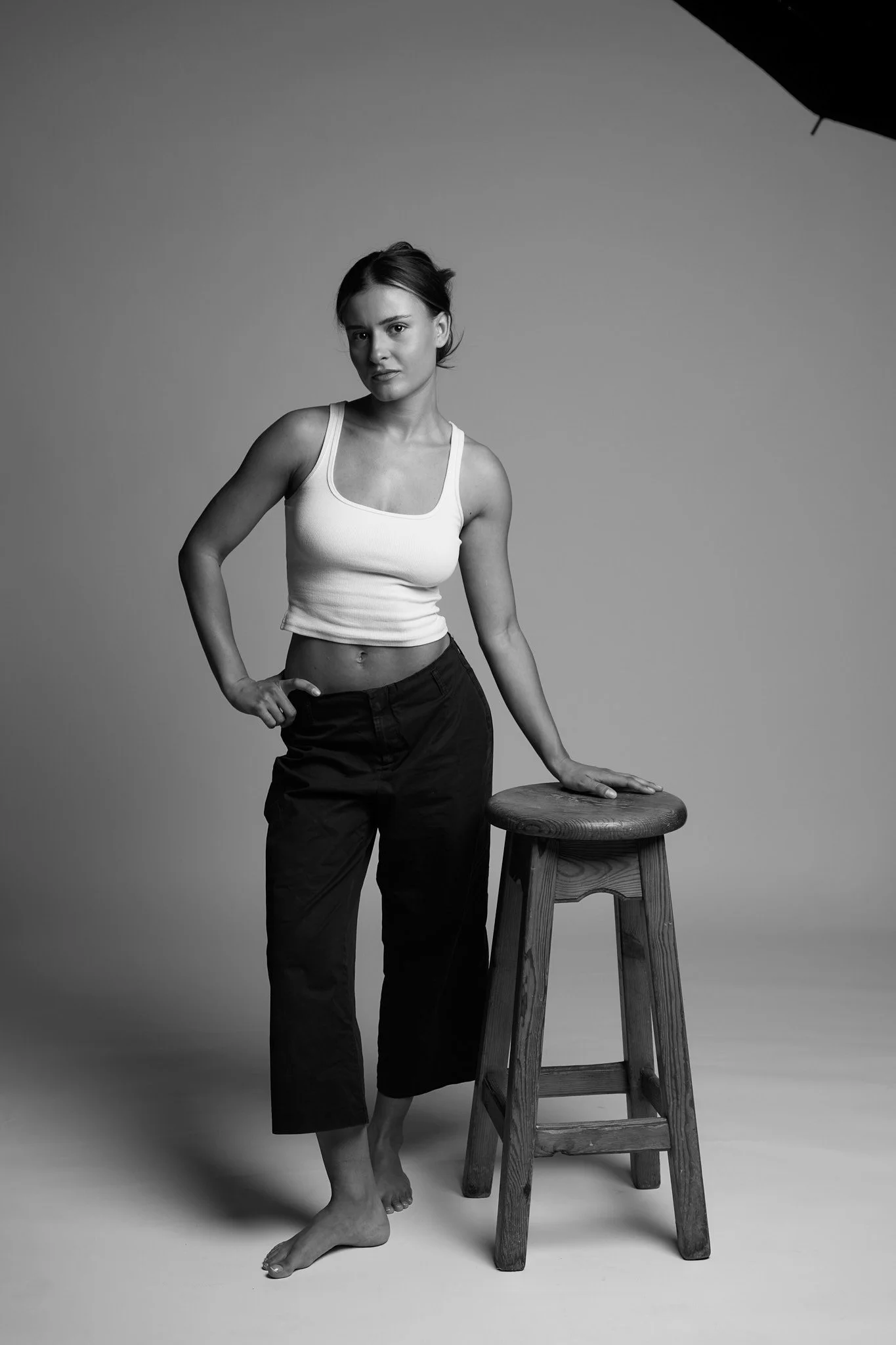A black and white photo of a woman standing next to a wooden stool. She is wearing a tank top and loose pants, with one hand on her hip and the other resting on the stool. She has short hair and a serious expression.