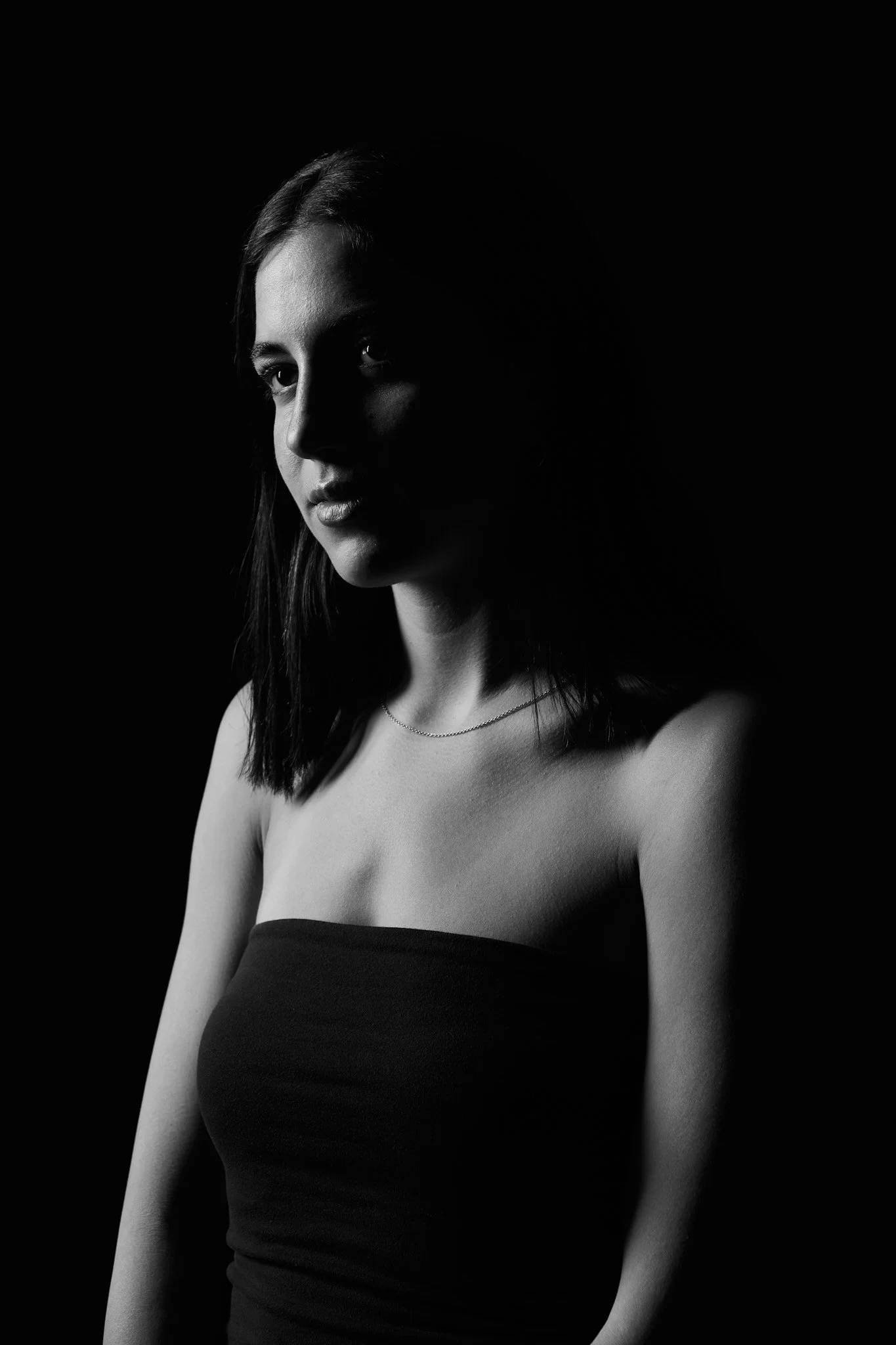 A black and white portrait of a young woman with shoulder-length hair, wearing a strapless top and a delicate necklace. She is looking slightly to the right, with a serious expression, contrasting with a dark background.