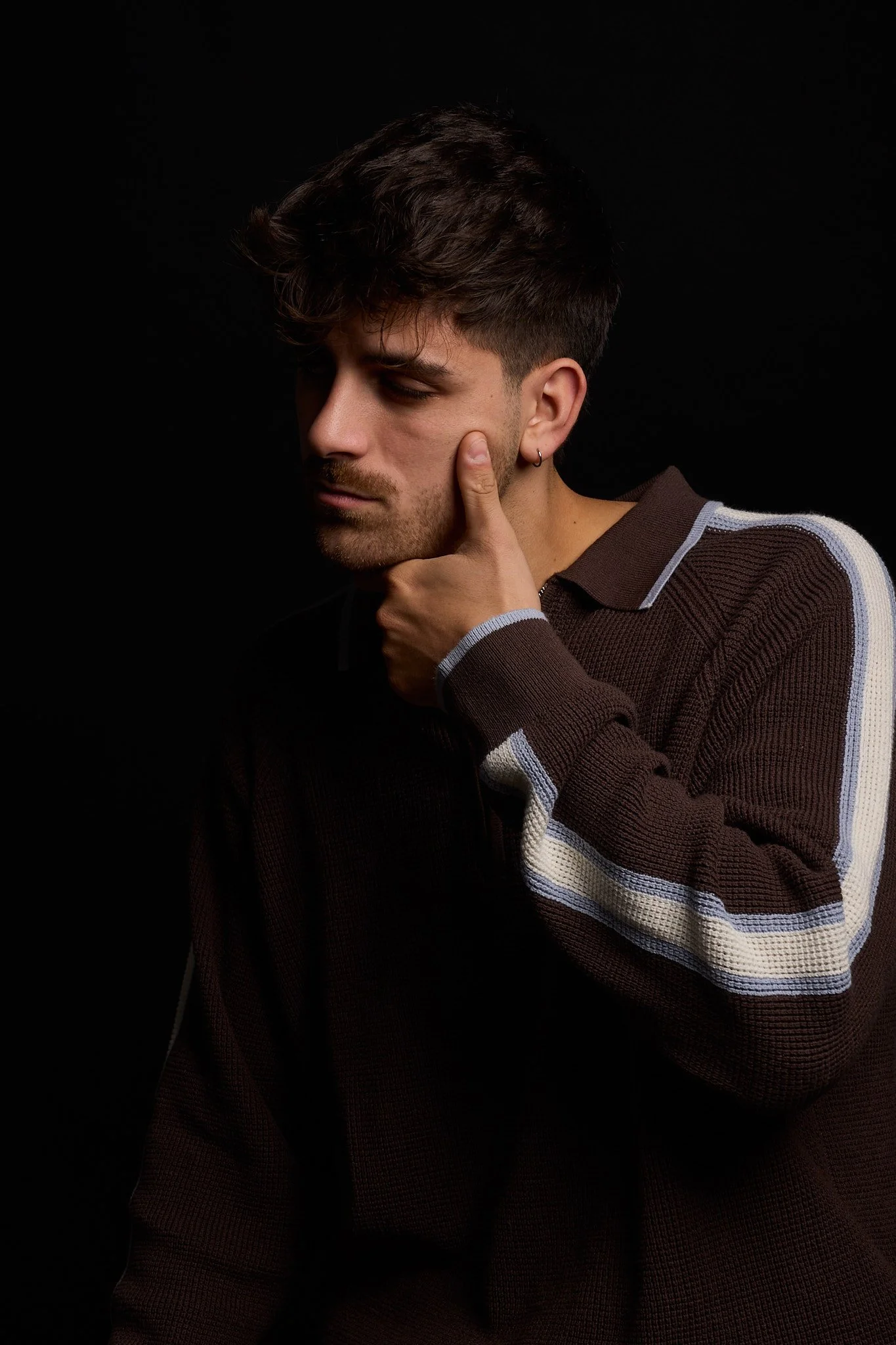 A young man with dark, wavy hair and a beard, wearing a brown sweater with white and gray stripes on the sleeves, is touching his face with his hand against a black background.
