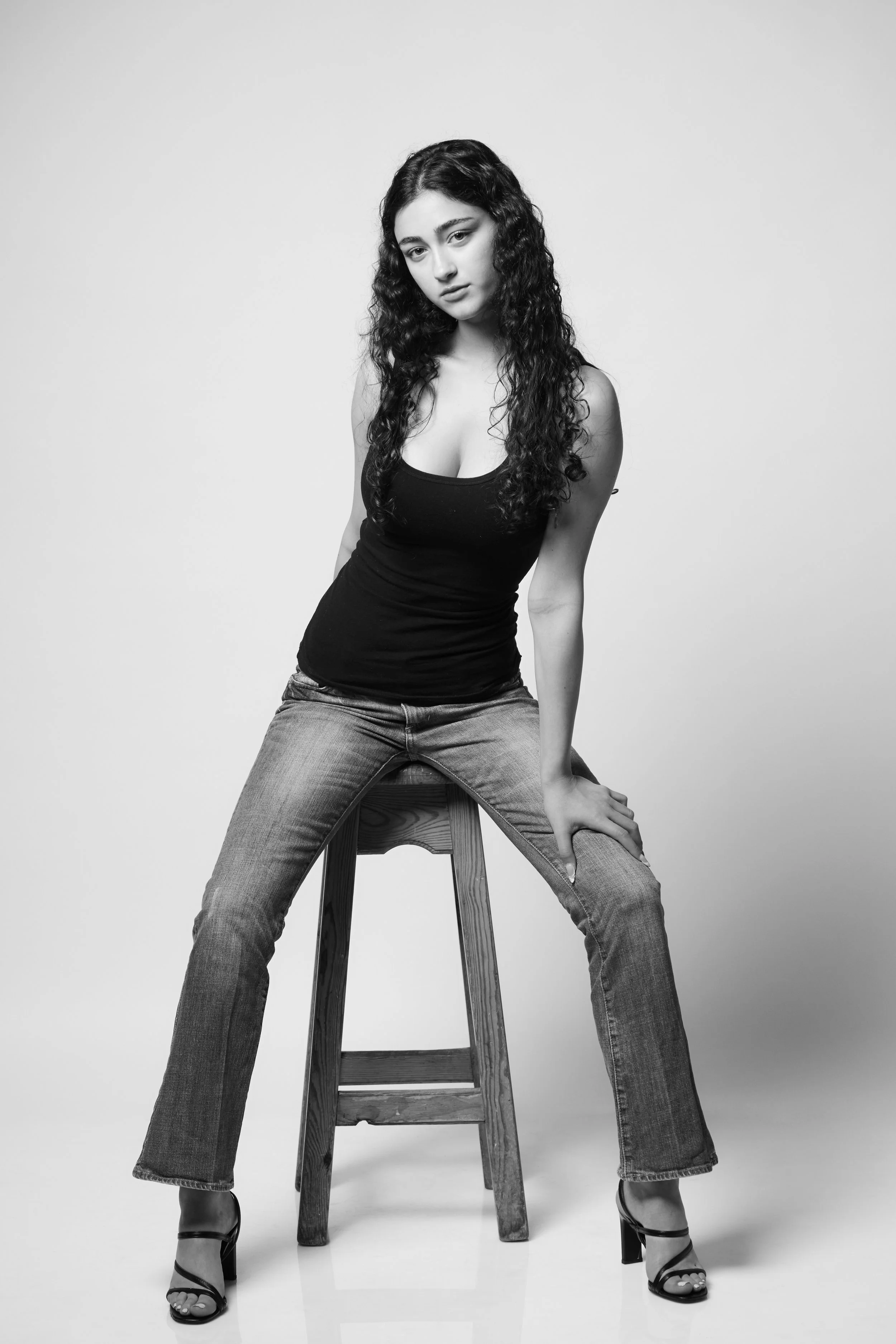 Black and white photo of a young woman with long, curly hair, wearing a tank top and jeans, sitting on a wooden stool, with her legs apart and hands resting on her thighs, in a studio setting with a plain background.