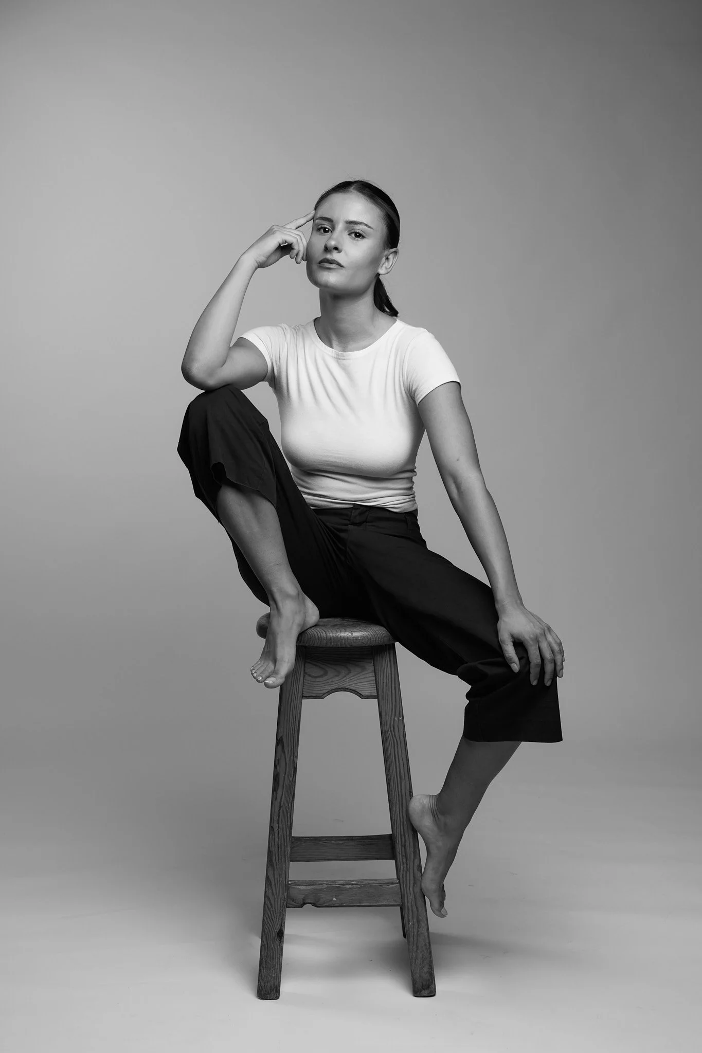 Black and white photo of a woman in a white t-shirt and dark pants, sitting on a stool with one foot on the ground and the other leg bent on the stool. She has her left hand resting on her knee and her right hand with her fingers touching her temple,