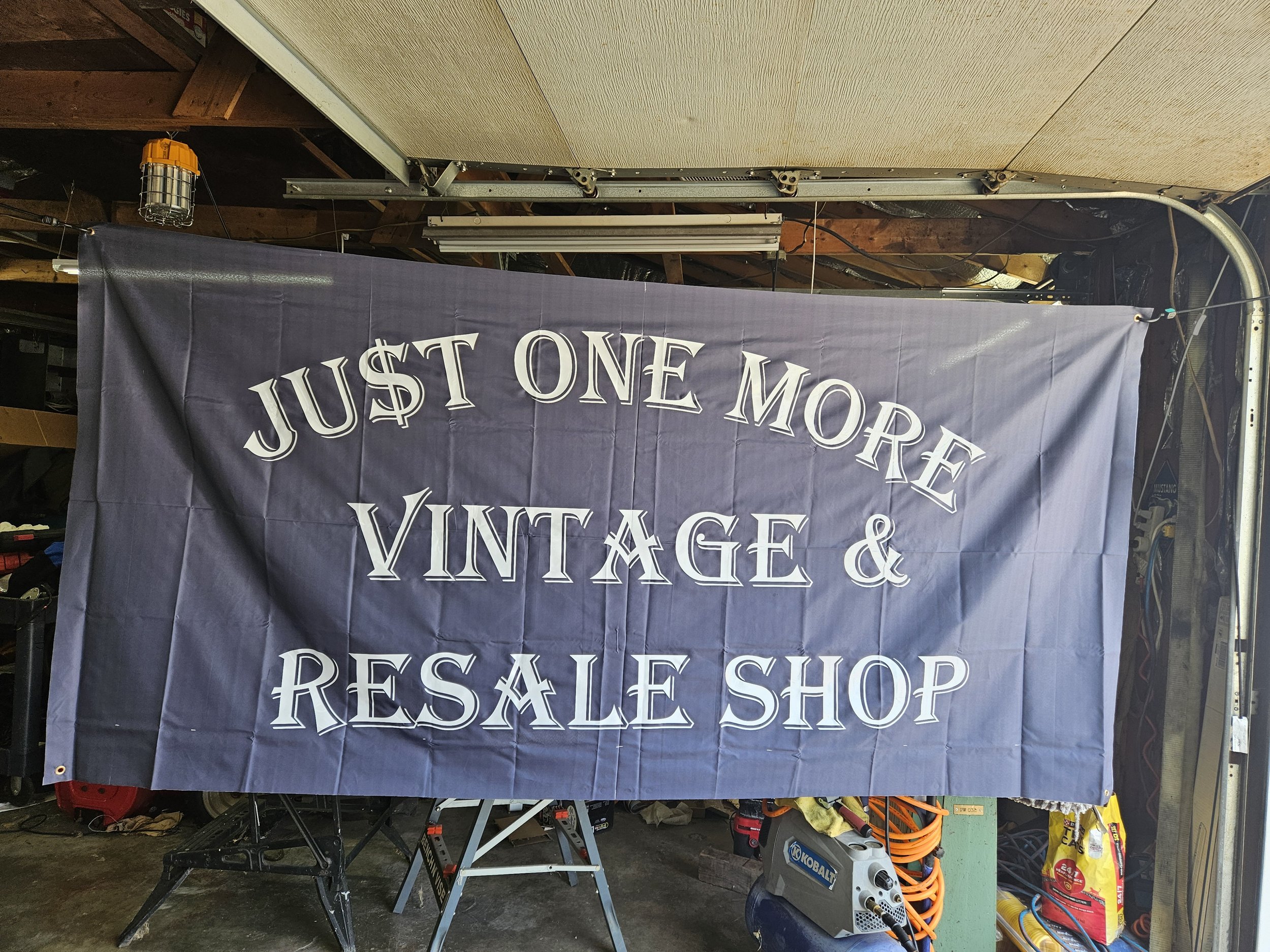 A large banner hanging in a garage or workshop, with the words "JUST ONE MORE VINTAGE & RESALE SHOP" written on it. Part of the garage ceiling and various tools and equipment are visible.