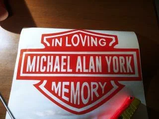 A shield-shaped sign with red and white colors, reading 'In Loving Memory of Michael Alan York.'