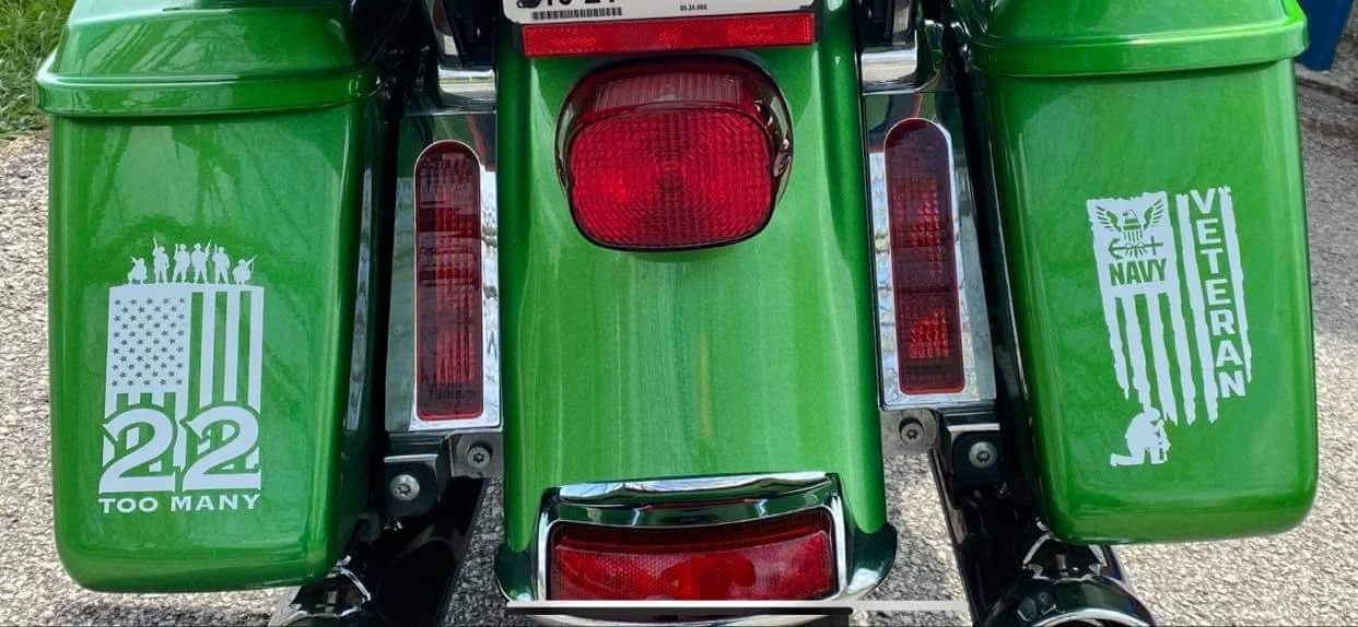 Rear view of a green motorcycle with two green saddlebags displaying patriotic stickers supporting veterans, the United States, and the Navy veterans.