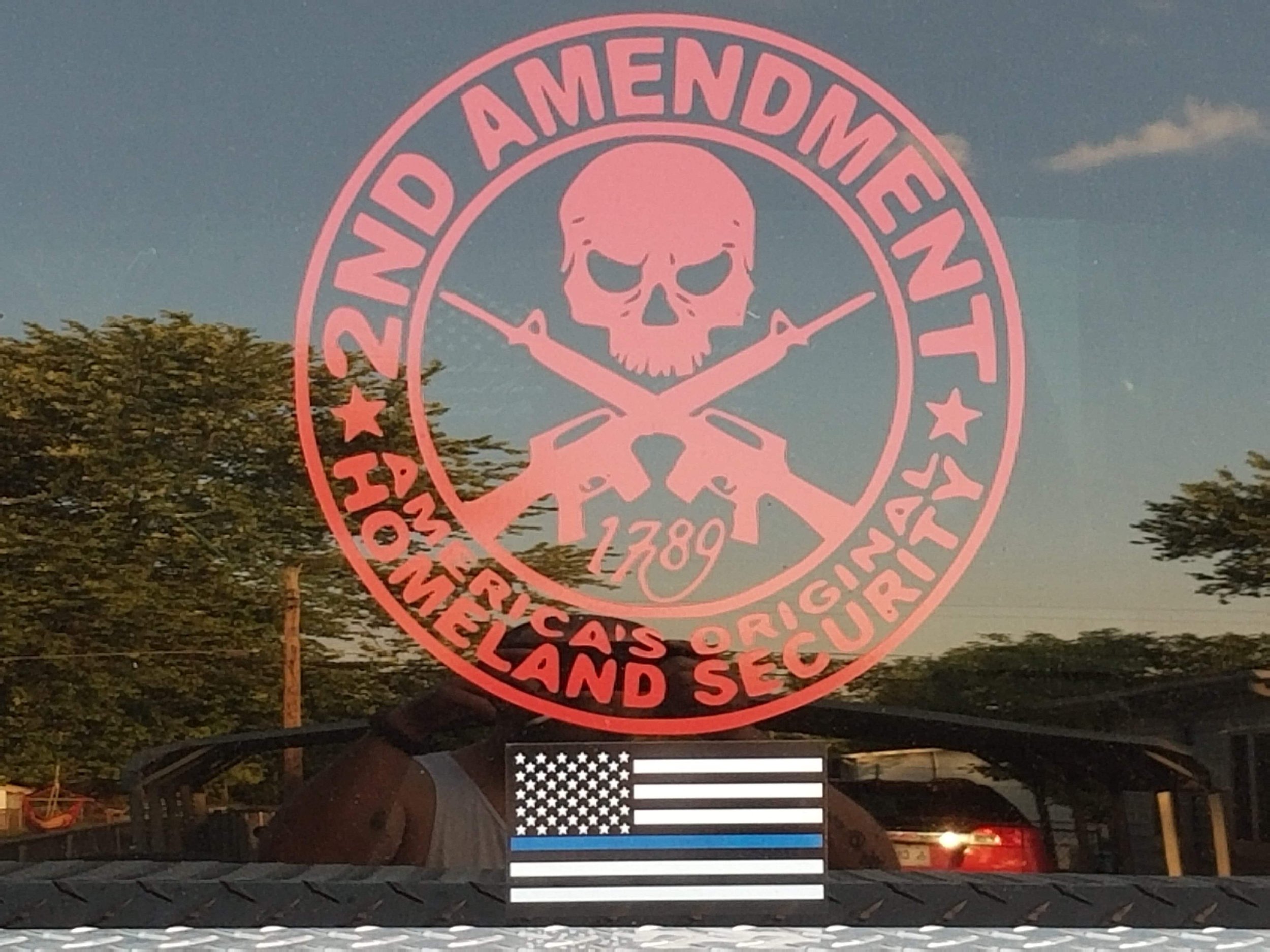 A large red circular sign with a skull and crossbones holding two crossed rifles and the words 'American Added Amendment' around the top. Below the sign, there is an American flag decal on a car windshield.