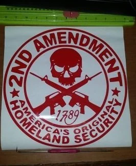 A circular sticker with a red and white design. It features a skull with a rifle crossing behind it, the text "2ND AMENDMENT" at the top, "HOMELAND SECURITY" at the bottom, and the year "1789" beneath the skull.