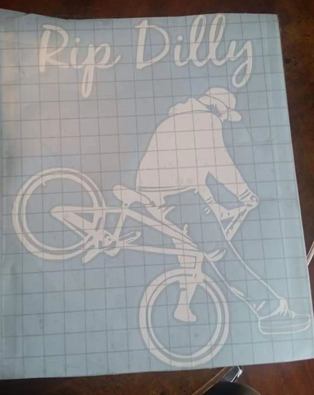 Design of a person riding a bicycle, wearing a hat, with the text 'Rip, Dilly' at the top, on grid paper.