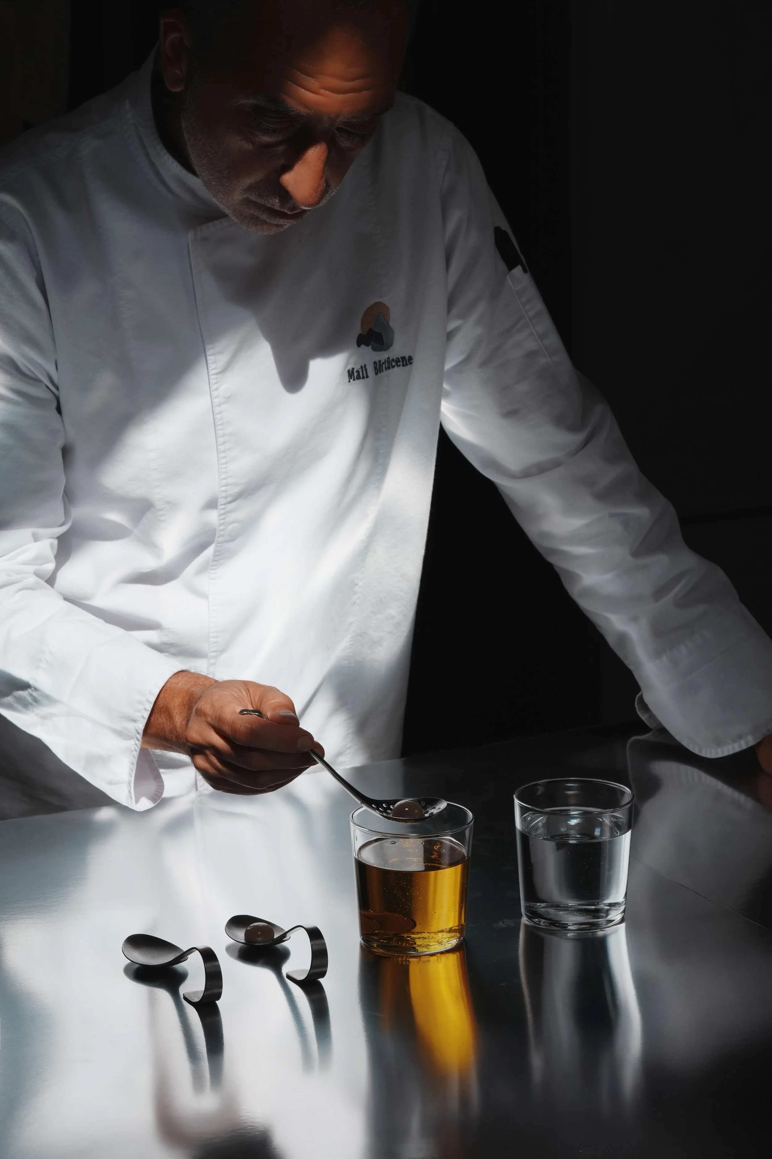 A chef with gray hair and a beard, dressed in a white chef's coat with his name "Mati Bolderone" embroidered on it, is carefully preparing food in a dimly lit kitchen. The surface in front of him holds two glass cups filled with dark liquid and two silver serving spoons.
