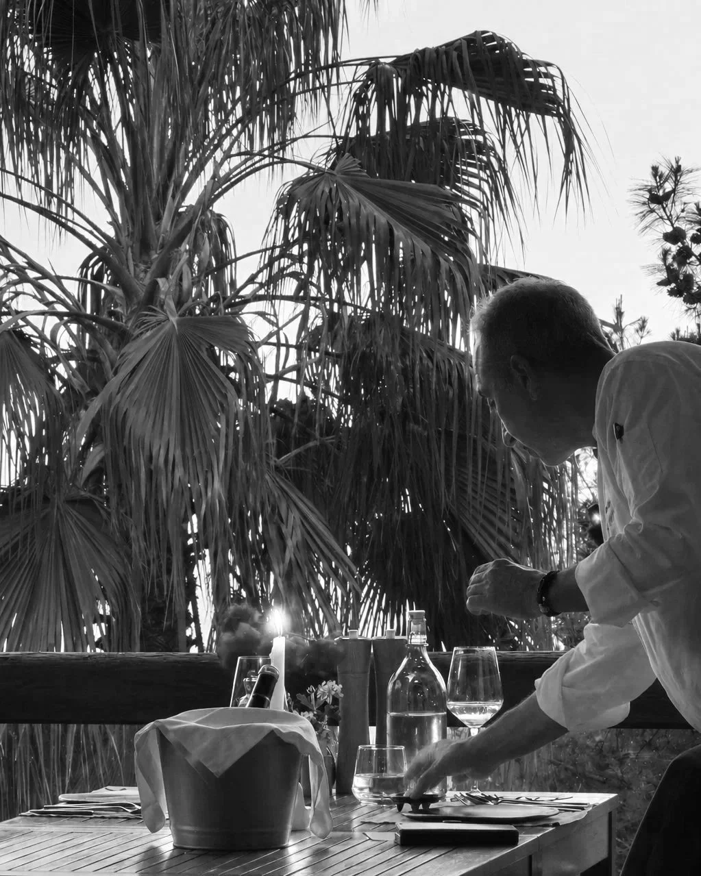 Chef Mehmet Ali Börtücene setting a table outdoors with trees and plants in the background, on a sunny day.