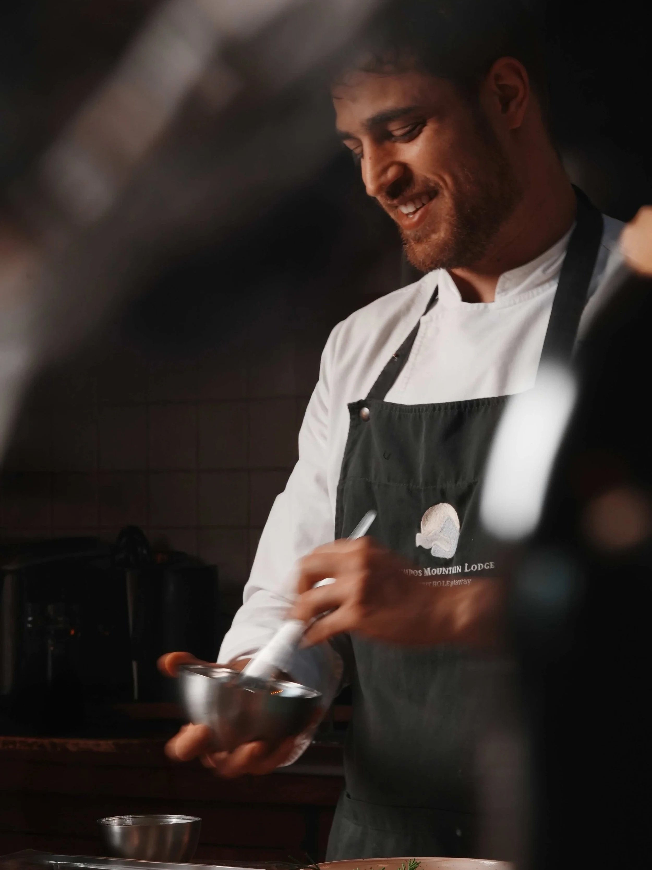 Close-up of sous-chef with brown hair and beard smiling, wearing a white shirt and a black apron, cooking or preparing food in a kitchen.