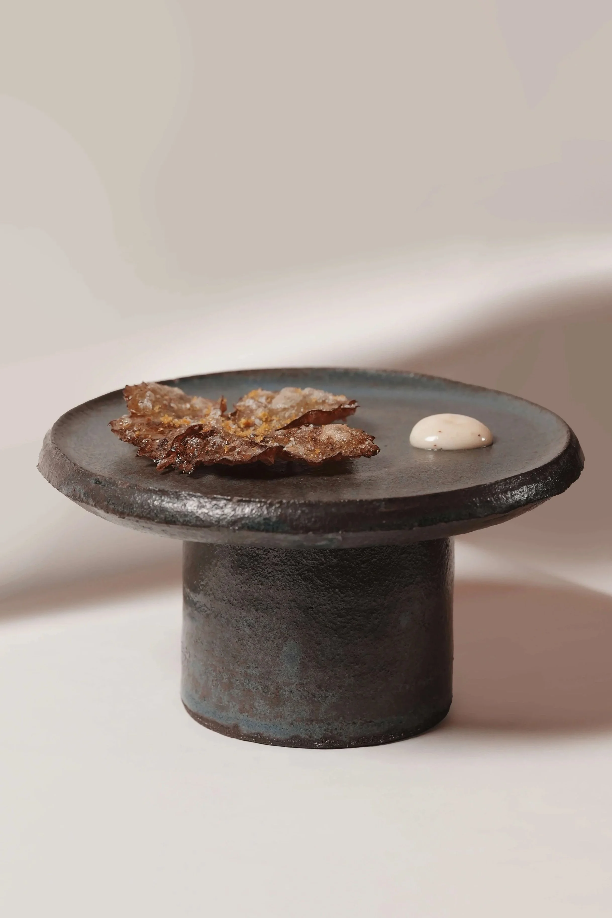 A black cylindrical stand with a flat, round top holding a piece of bark and a dollop of cream-colored sauce
