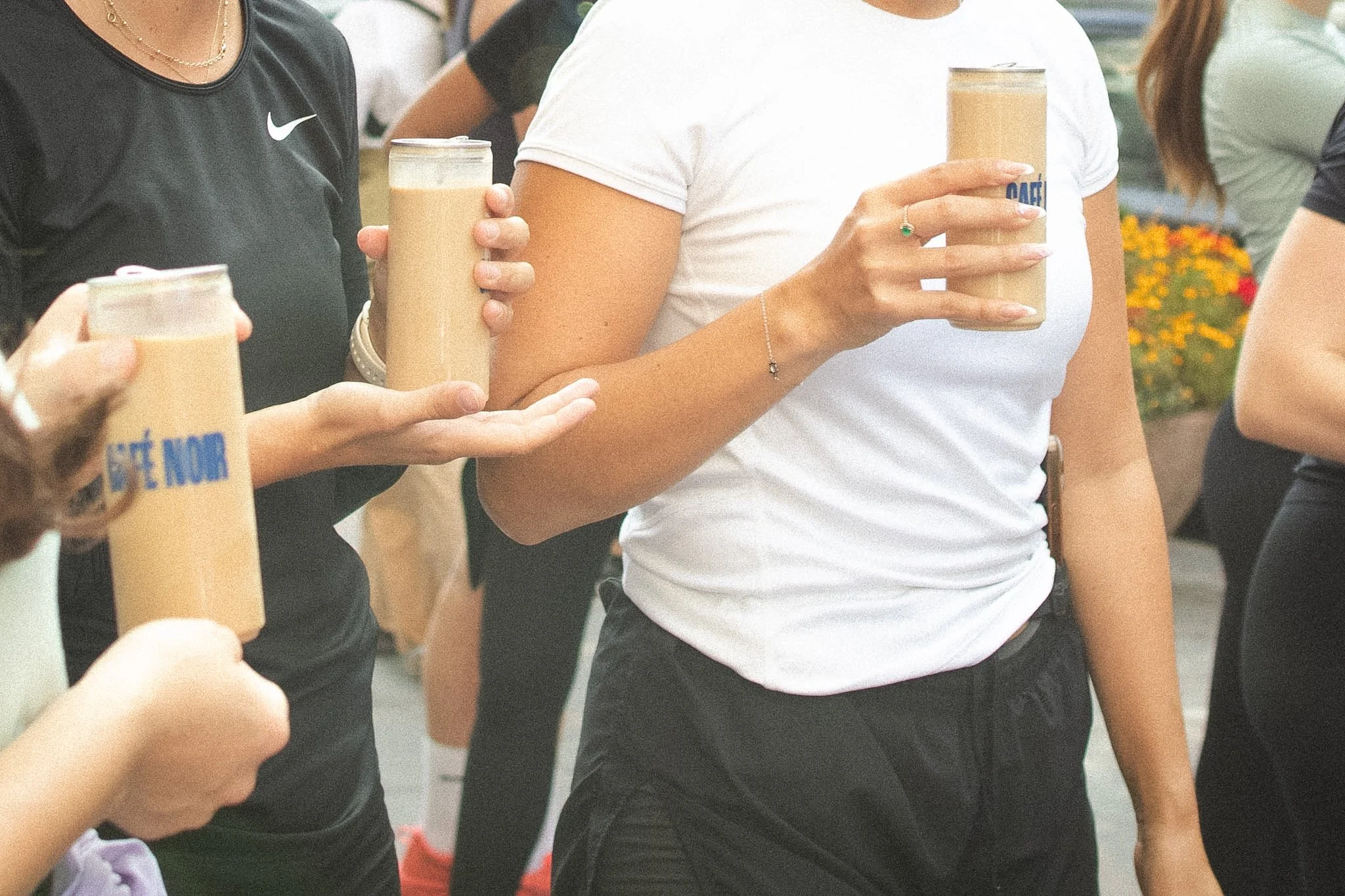 Group of people standing outdoors, holding drinks in beige cups, with some wearing athletic clothing and jewelry.