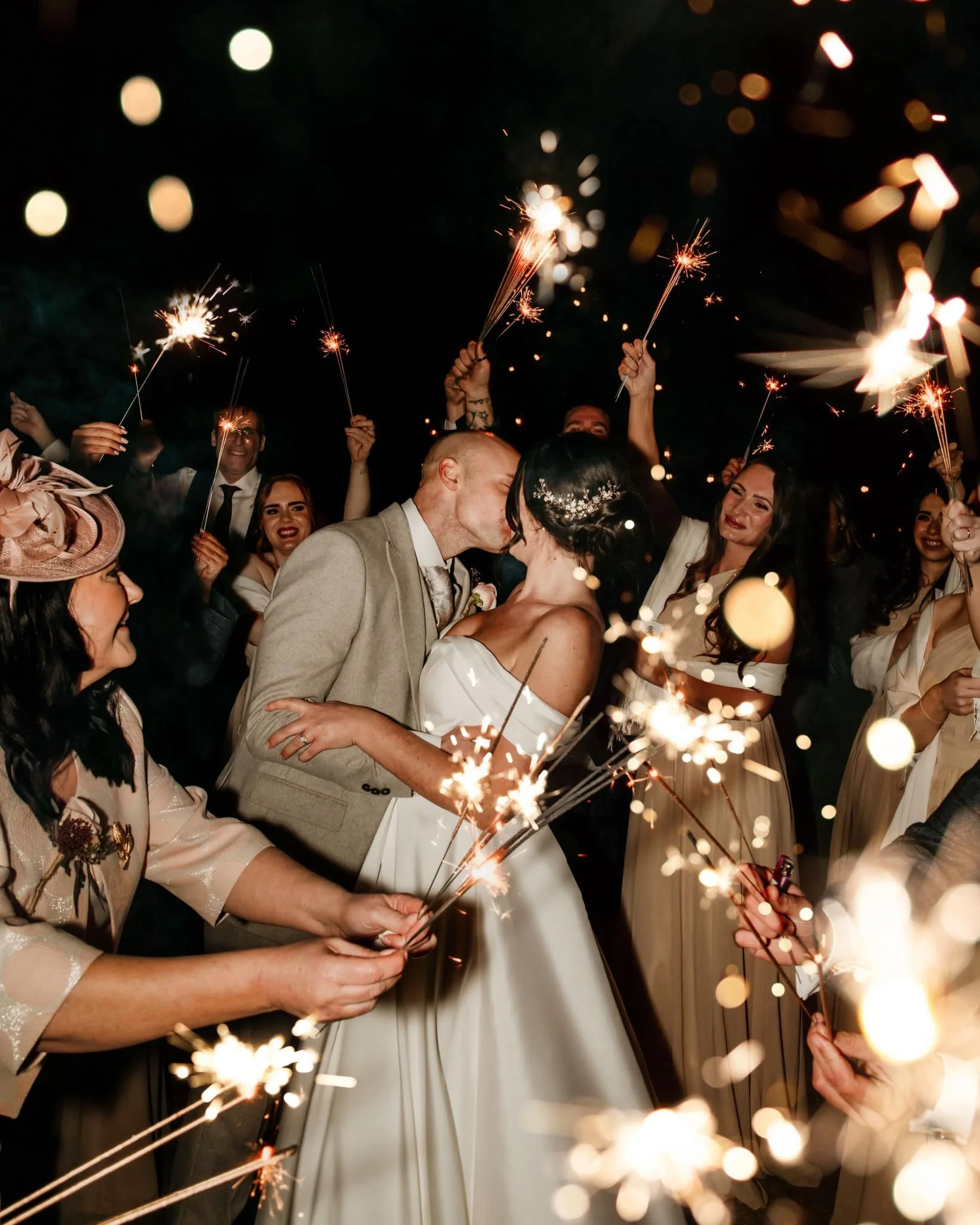 A group of people celebrating at night, holding sparklers and enjoying a wedding kiss, with smiles and festive mood.
