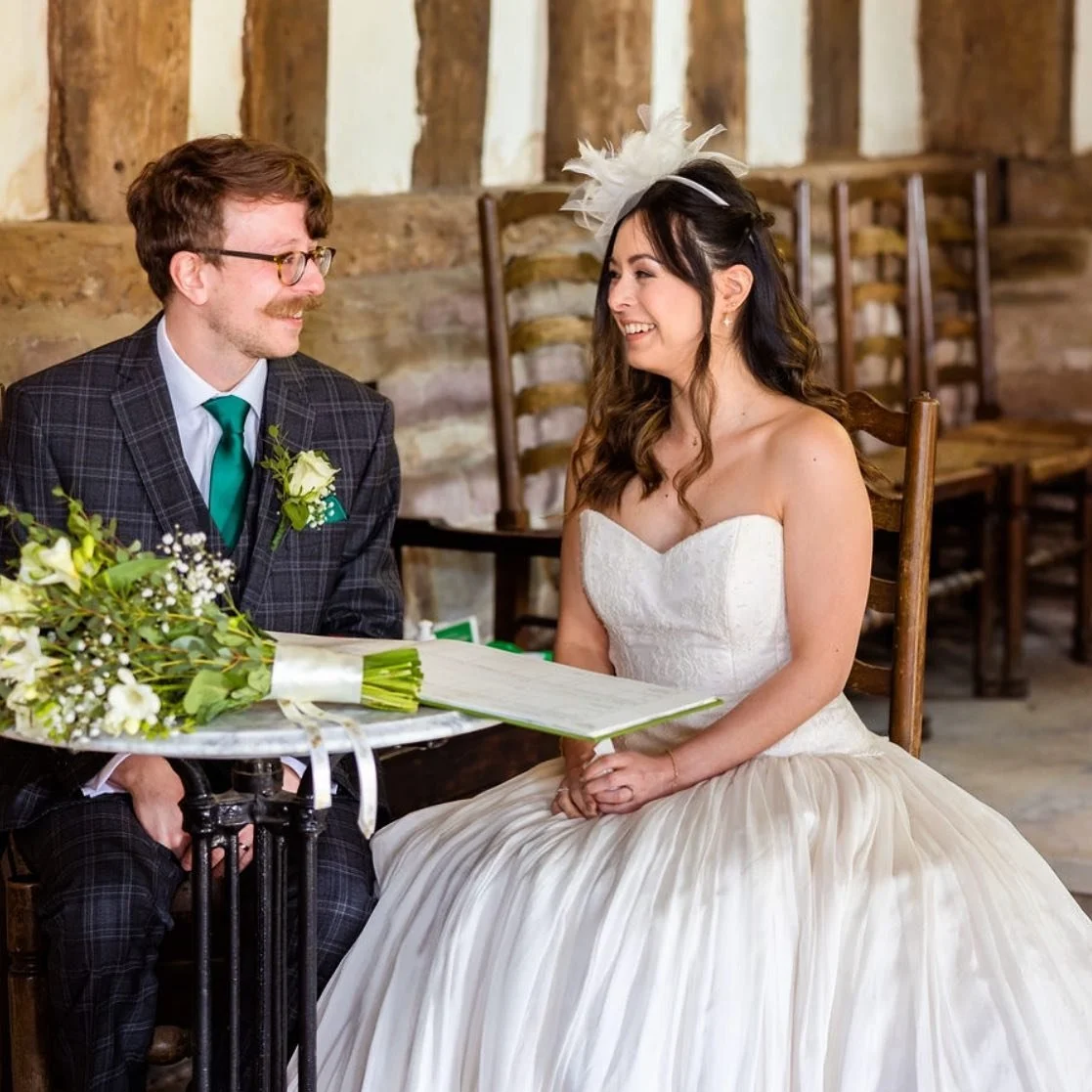 A bride and groom sitting at a wedding ceremony, smiling at each other. The groom wears glasses, a checkered suit with a green tie, and a white boutonniere. The bride wears a strapless white gown with a full skirt and a feathered headpiece. There is 