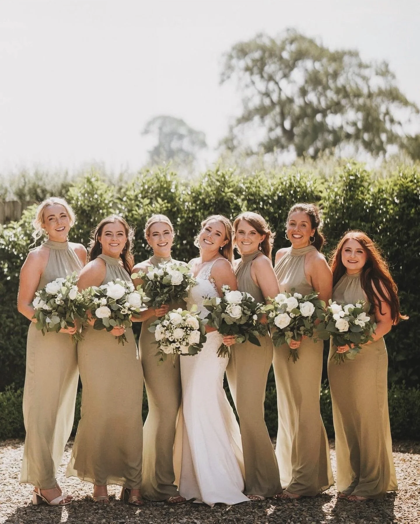 Bridal party with seven women, six bridesmaids in matching beige dresses, and a bride in a white wedding gown, standing outdoors with greenery and trees in the background, each holding a bouquet of white flowers.