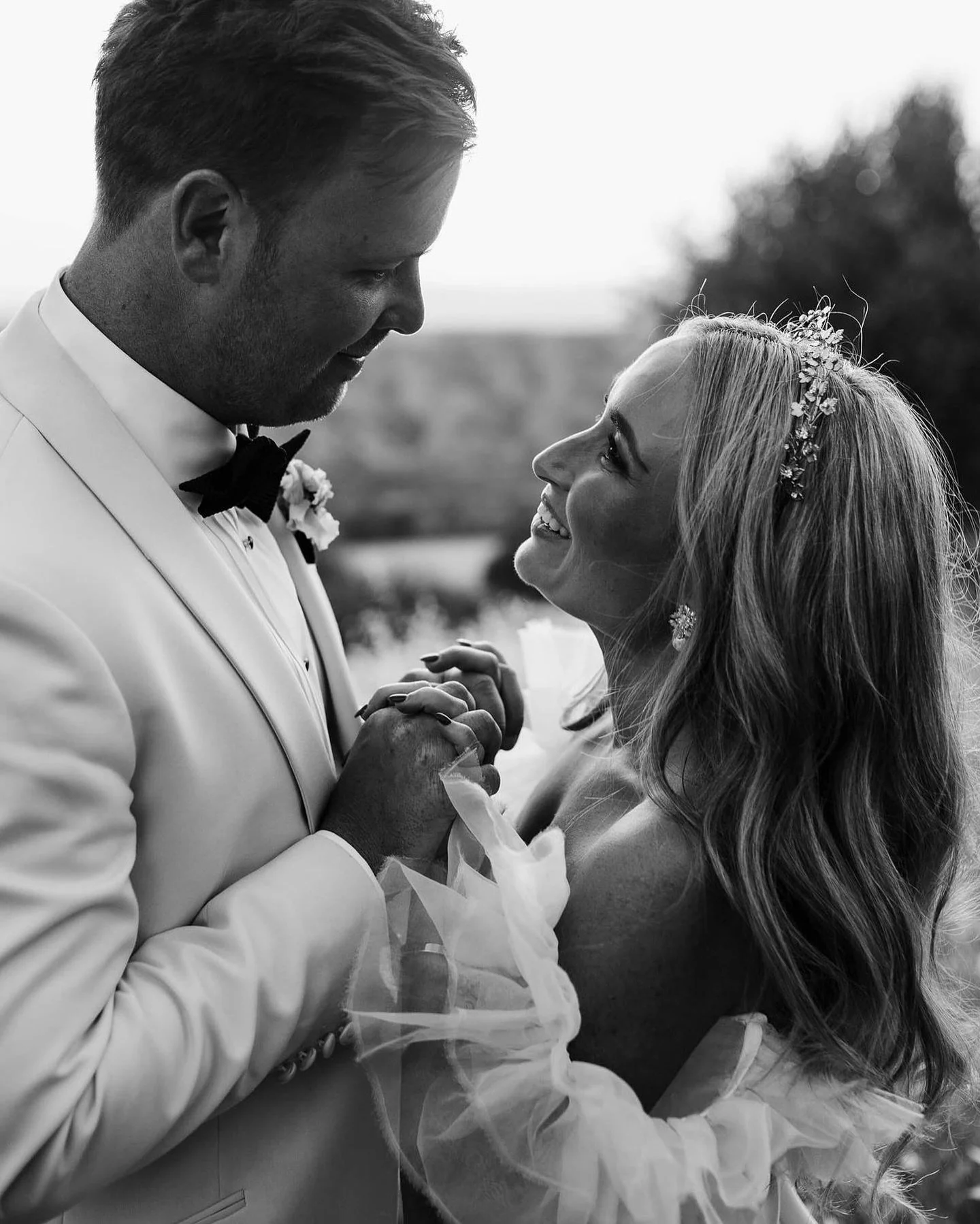 A black and white photo of a man and woman holding hands and gazing into each other's eyes, dressed in wedding attire, outdoors.