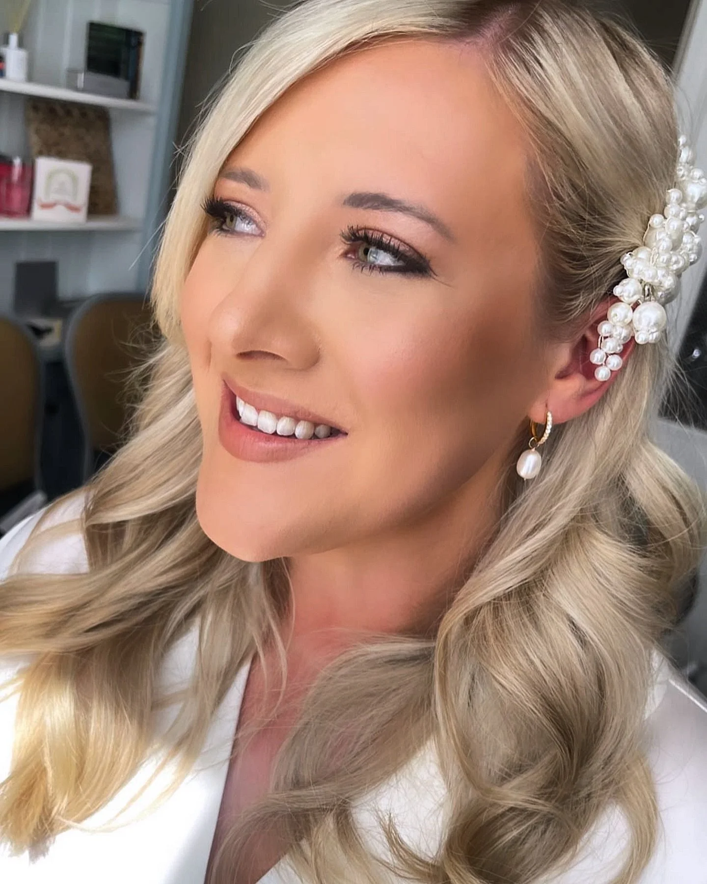 Close-up of a woman with long wavy blonde hair, wearing pearl jewelry, smiling and looking to the side in an indoor setting.
