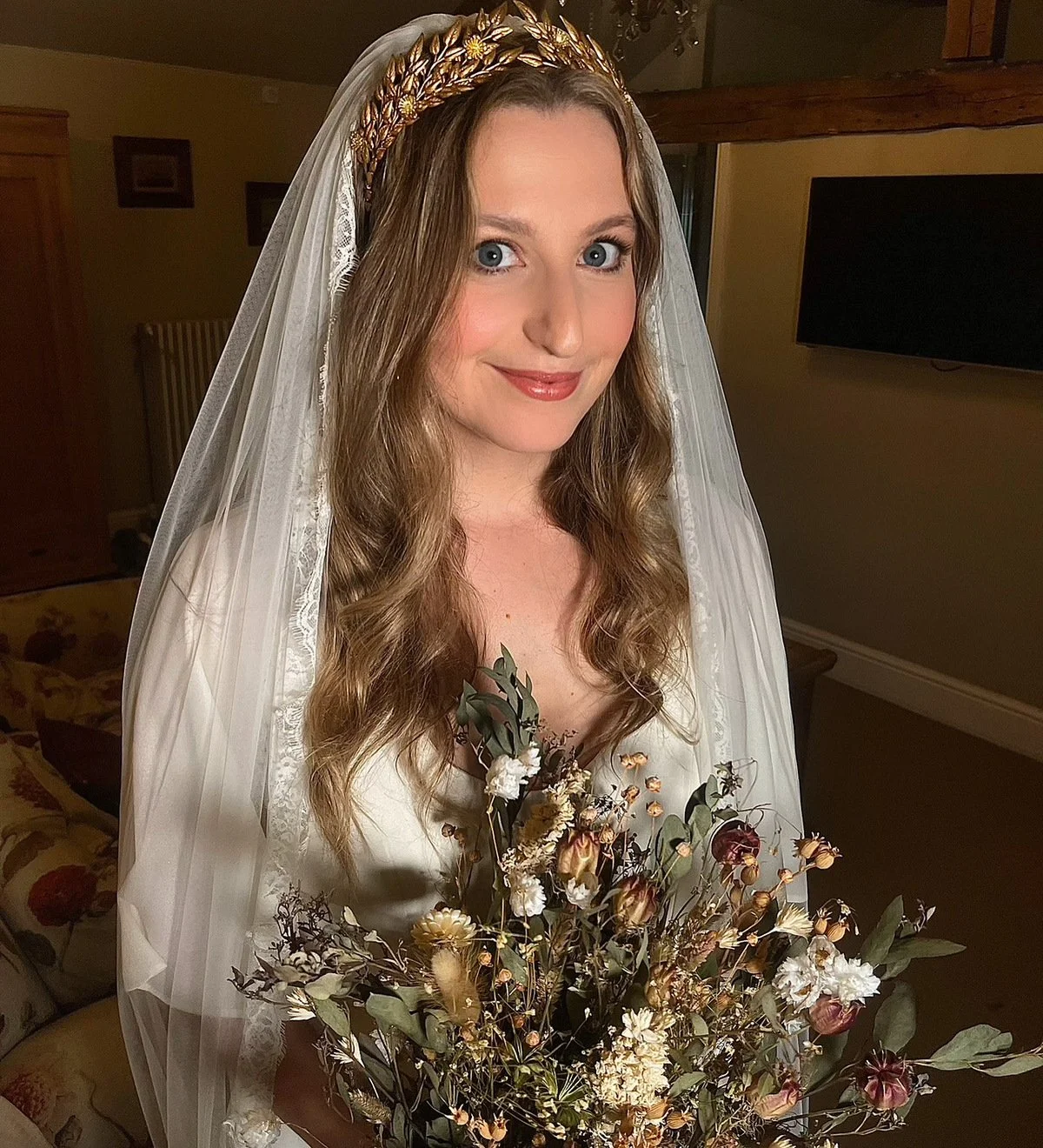A woman with long wavy hair, wearing a white wedding dress and veil, holding a bouquet of dried flowers, standing indoors in a warmly lit room.