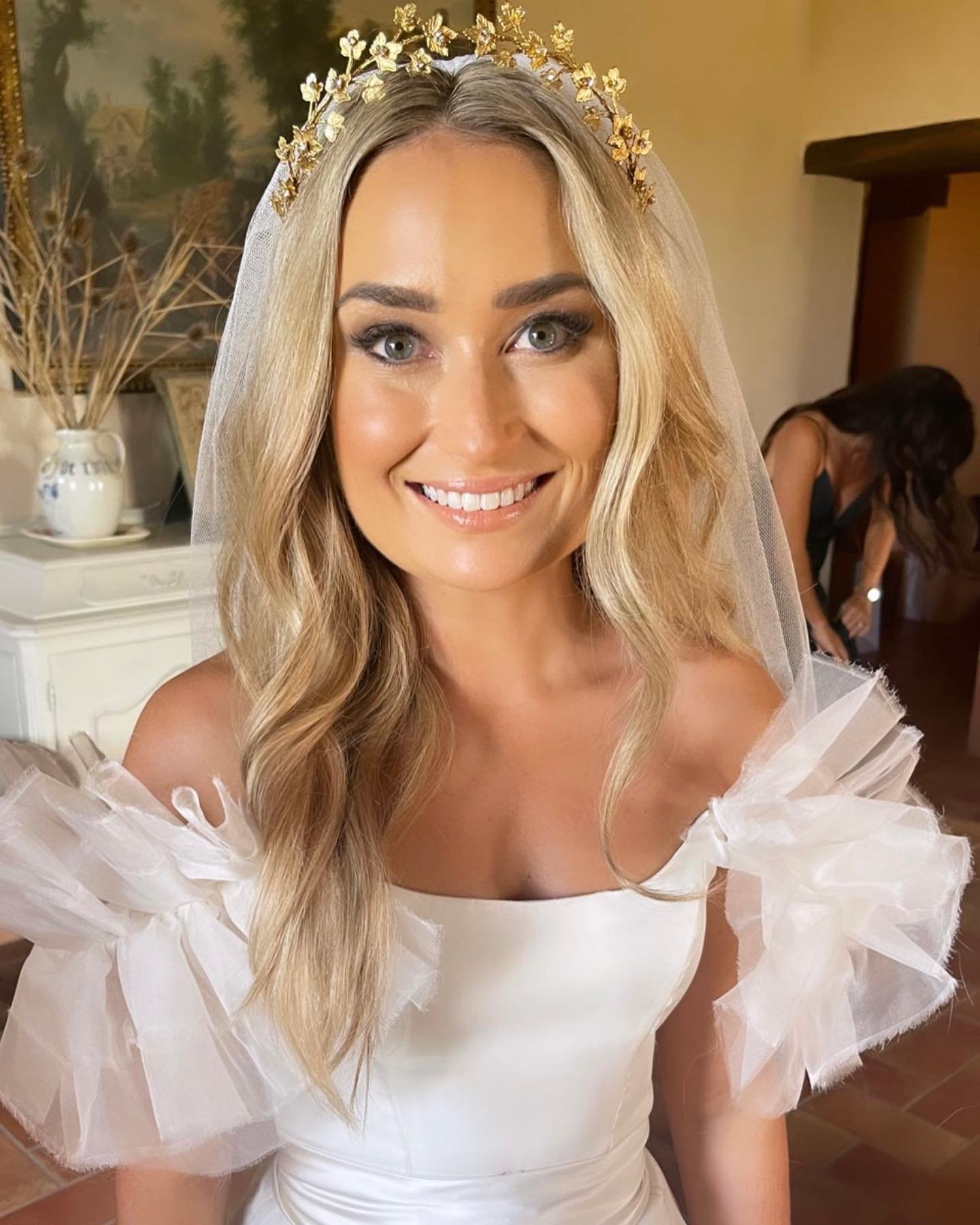 A smiling bride with long blonde hair, wearing a white dress with tulle puff sleeves, a gold butterfly headpiece, and a veil. She is indoors with a woman in black in the background.