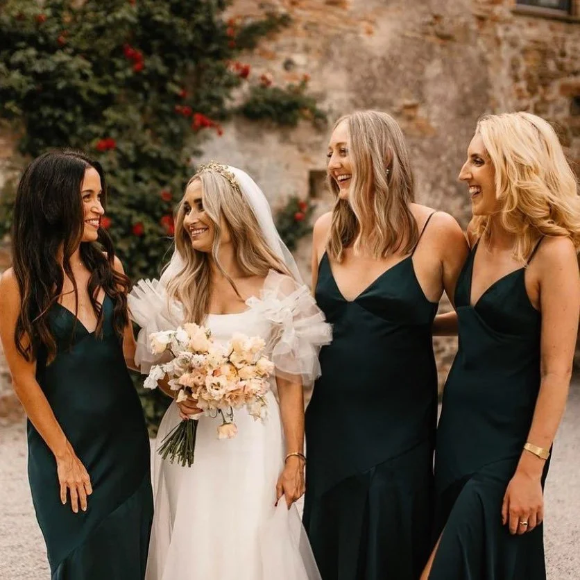 Four women standing together at a wedding, smiling and talking. The bride is holding a bouquet of flowers and wearing a white wedding dress with ruffled sleeves and a veil. The other three women are in matching dark green dresses.