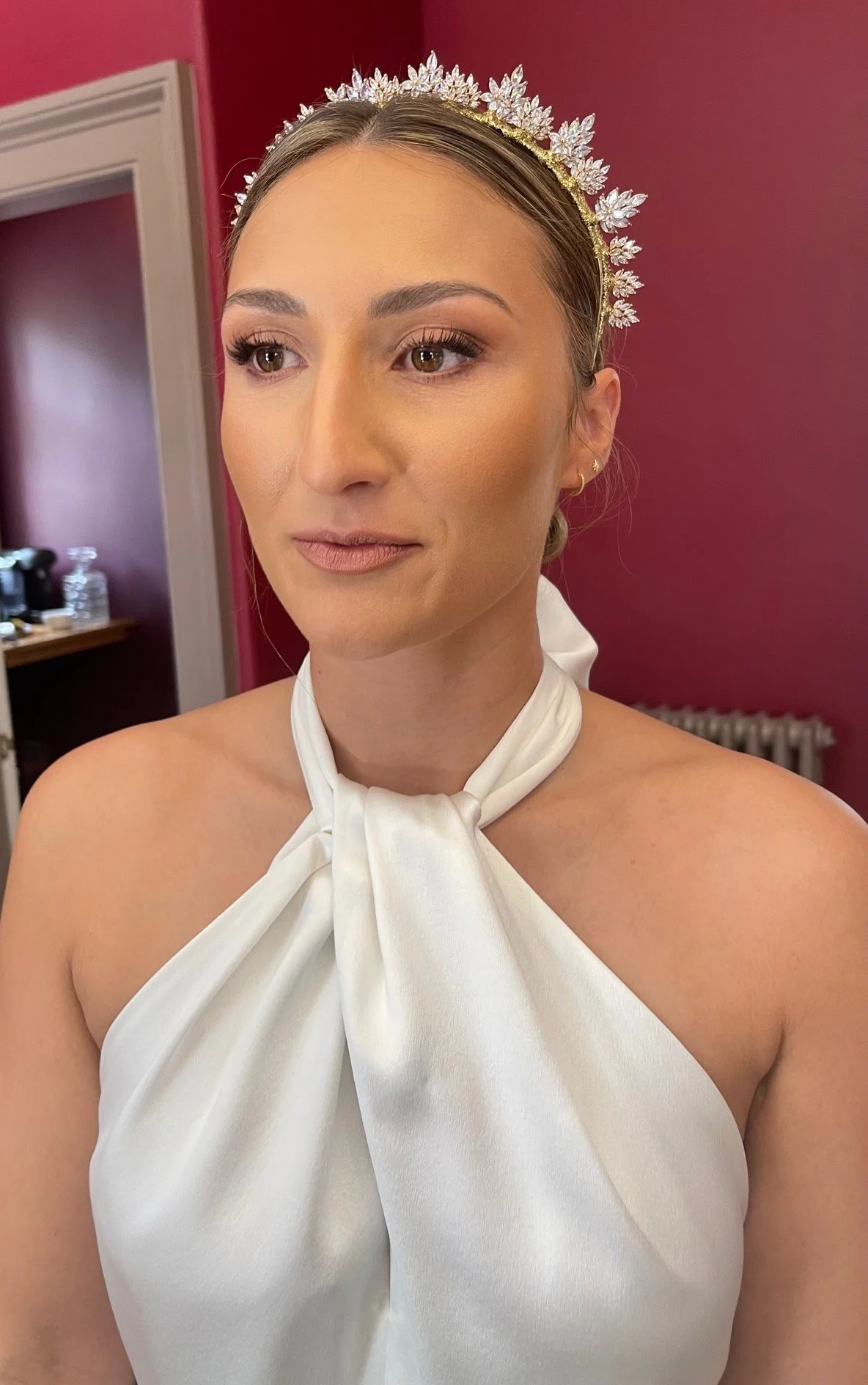A woman with brown hair wearing a white satin dress with a knot at the neck, a jeweled leaf crown, and small hoop earrings, standing in a room with pink and red walls.