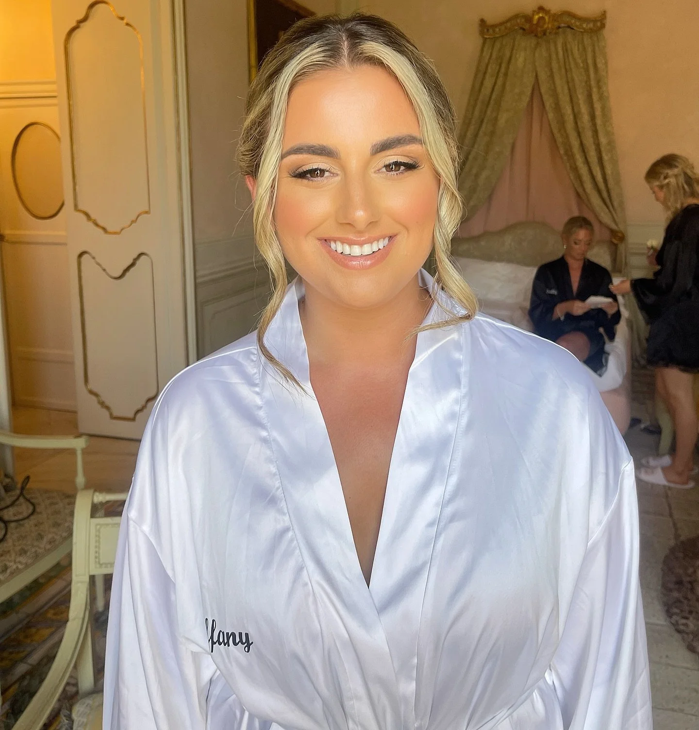 A smiling woman with blonde hair styled in loose waves, wearing a white satin robe, standing in a luxurious bedroom with ornate decor and drapery, and other women in the background preparing.