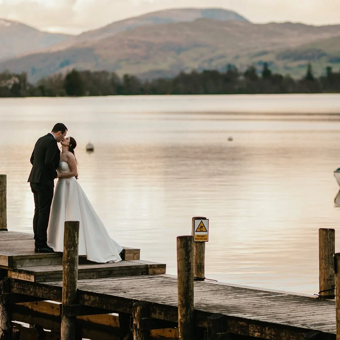 A couple in wedding attire sharing a kiss on a wooden dock by the water, with mountains in the background at sunset.