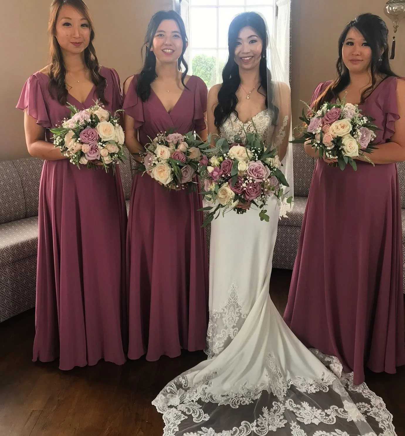 A bride and three bridesmaids at a wedding. The bride wears a white dress with lace detail and a long train, holding a large bouquet of pink, purple, and white flowers. The bridesmaids wear matching mauve dresses with ruffled sleeves, each holding a 