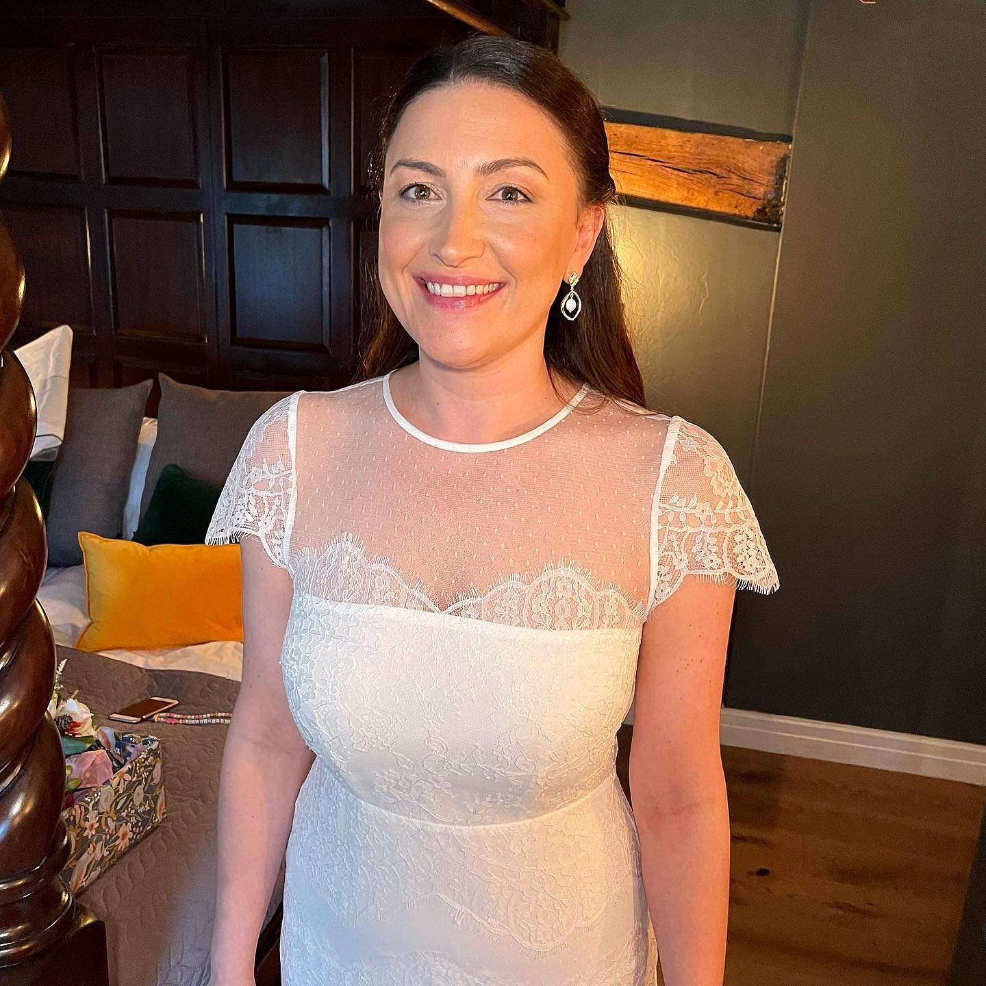 Woman in a white lace dress smiling indoors, with a dark wood headboard and colorful pillows on a bed in the background.