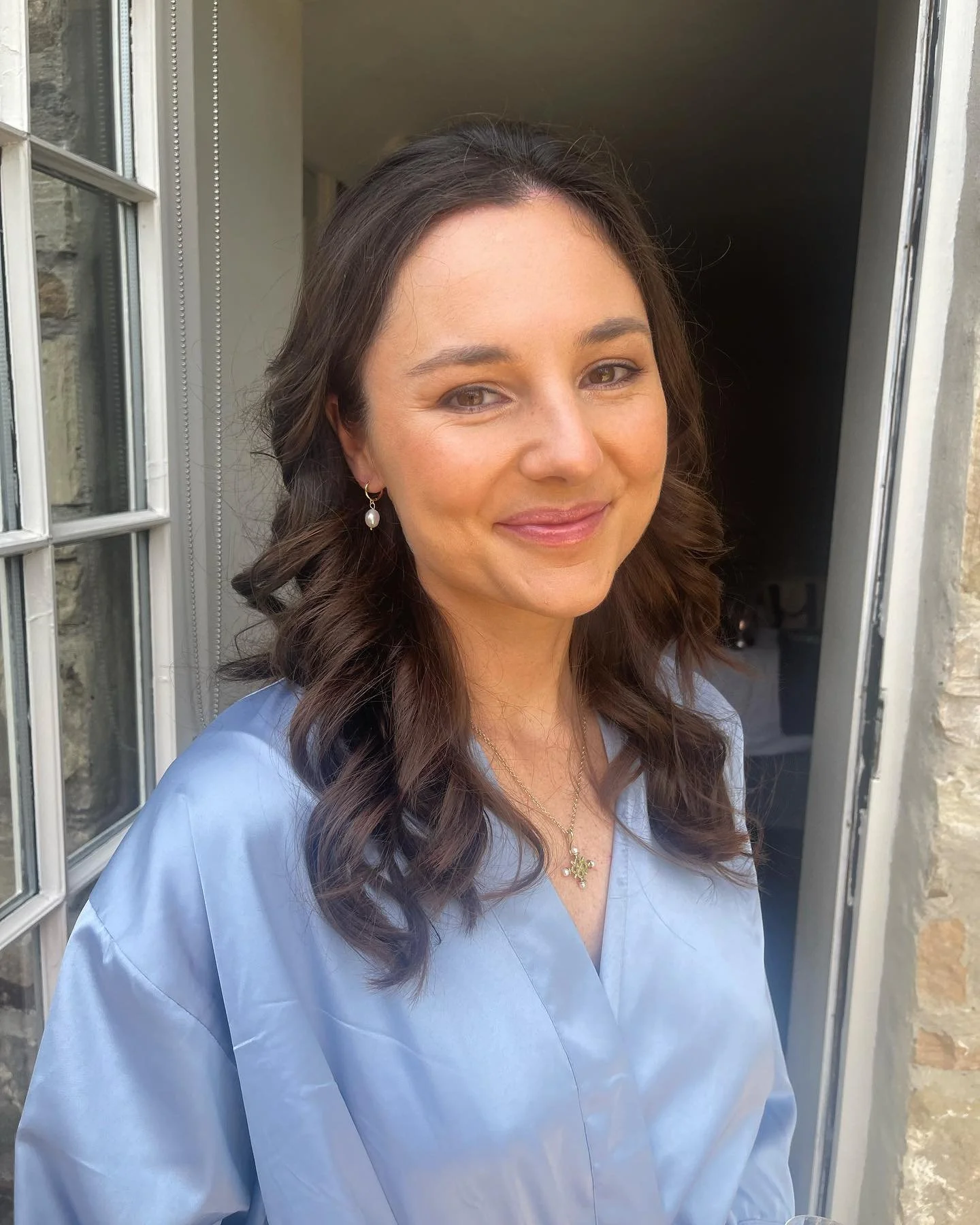 A woman with long curly brown hair wearing a light blue scrub top, pearl earrings, and a necklace with a cross pendant, standing in a doorway outdoors, smiling at the camera.