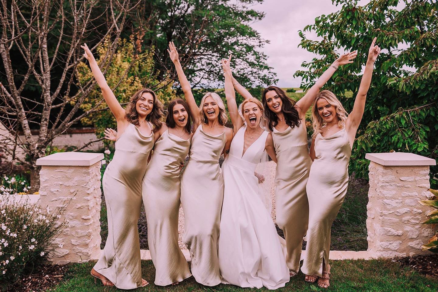 Group of six women dressed in elegant satin dresses, posing outdoors with arms raised and smiling, during a celebration or wedding, with lush greenery and stone pillars in the background.